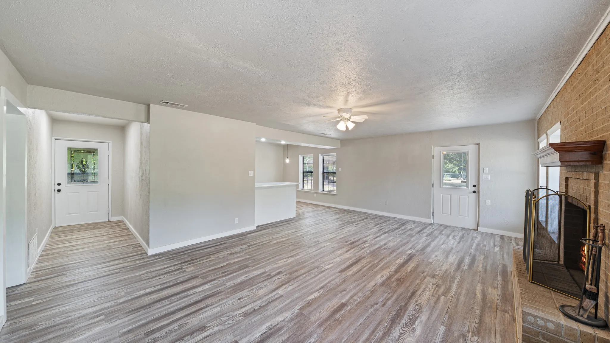 Spacious interior featuring wood-finish flooring, a brick fireplace with a wood mantel, a ceiling fan, and multiple windows for natural illumination