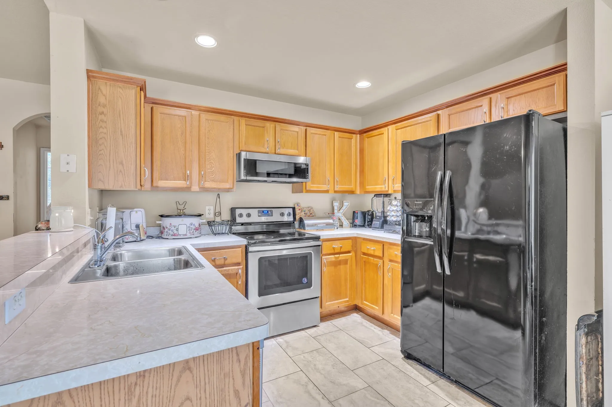 Kitchen featuring wood cabinetry, light-toned countertops, a stainless steel range with overhead microwave, a black side-by-side refrigerator, and tile flooring