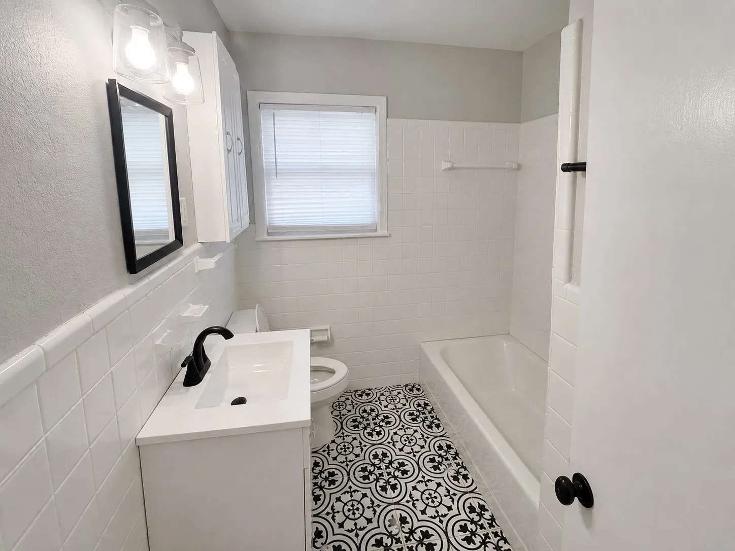 White subway tile bath surround, patterned floor tile, white vanity with integrated sink, black-framed mirror, and a white medicine cabinet
