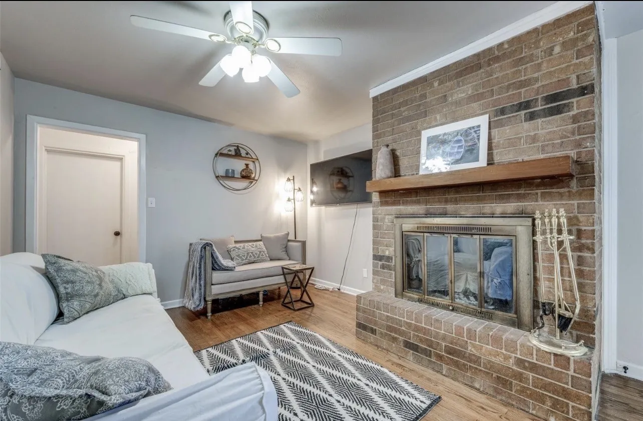 Brick-clad fireplace with a wood mantel, wood-finish flooring, and a ceiling fan