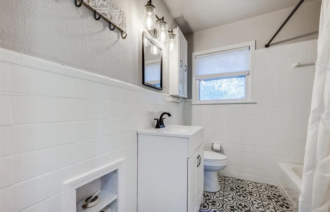 Patterned floor tile, white vanity with integrated sink, wall-mounted medicine cabinet, white subway tile wainscoting, and a window with a privacy blind