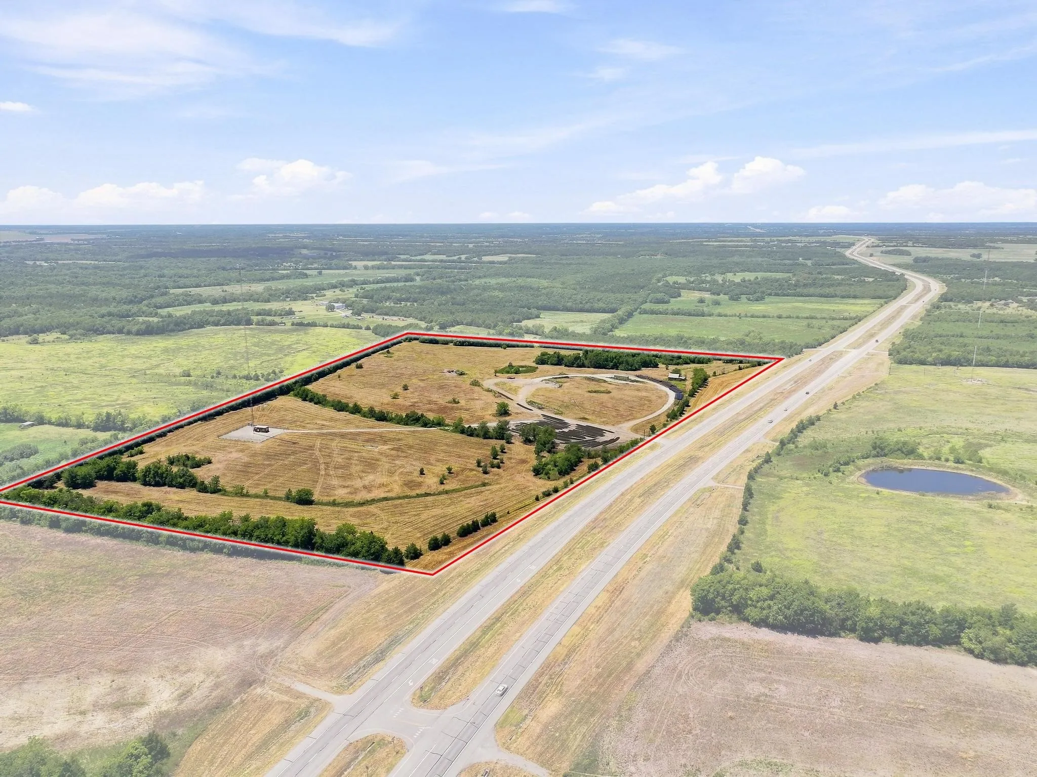 Expansive cleared land parcel bordered by mature trees, featuring a visible roadway, and a small pond within the property lines