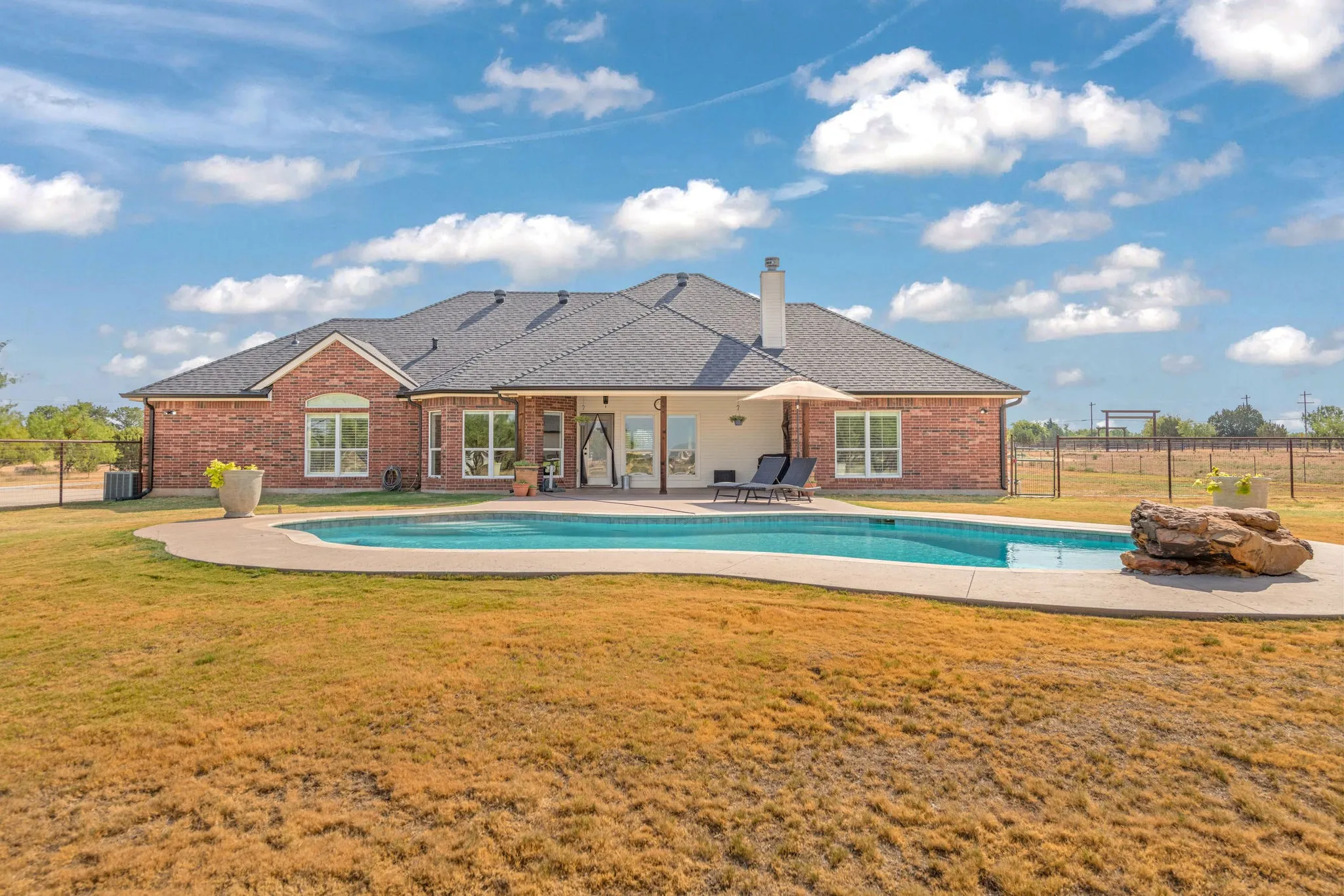 Brick exterior with a dark shingle roof and a covered back porch.  In ground swimming pool with jumping stone.