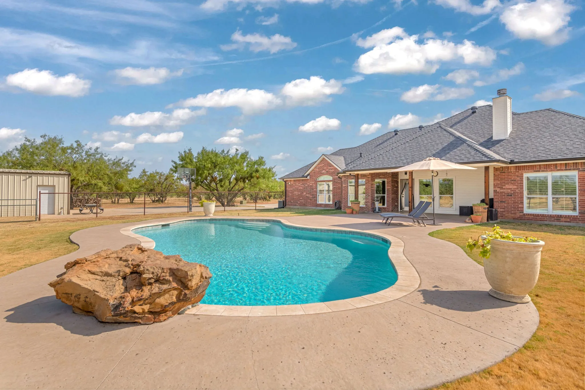 Freeform swimming pool with a surrounding concrete patio, brick home, and a detached metal workshop.