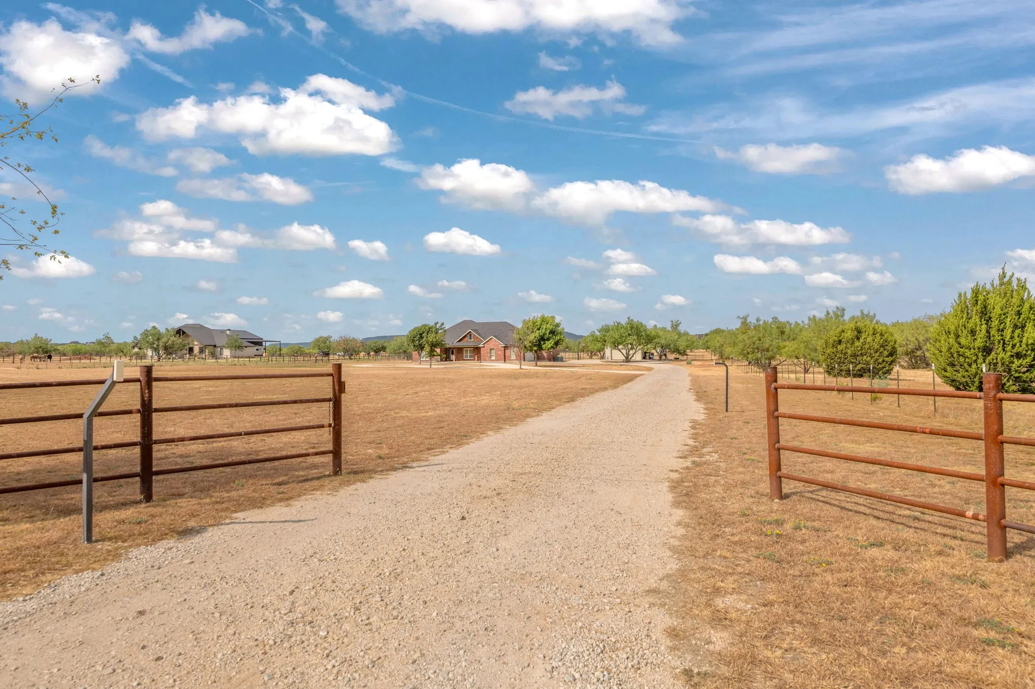 Gravel driveway bordered by pipe fenced entry, leading to a brick-clad residence with a dark Class 4 shingled roof.  T post fencing.  Room to roam on five acres.