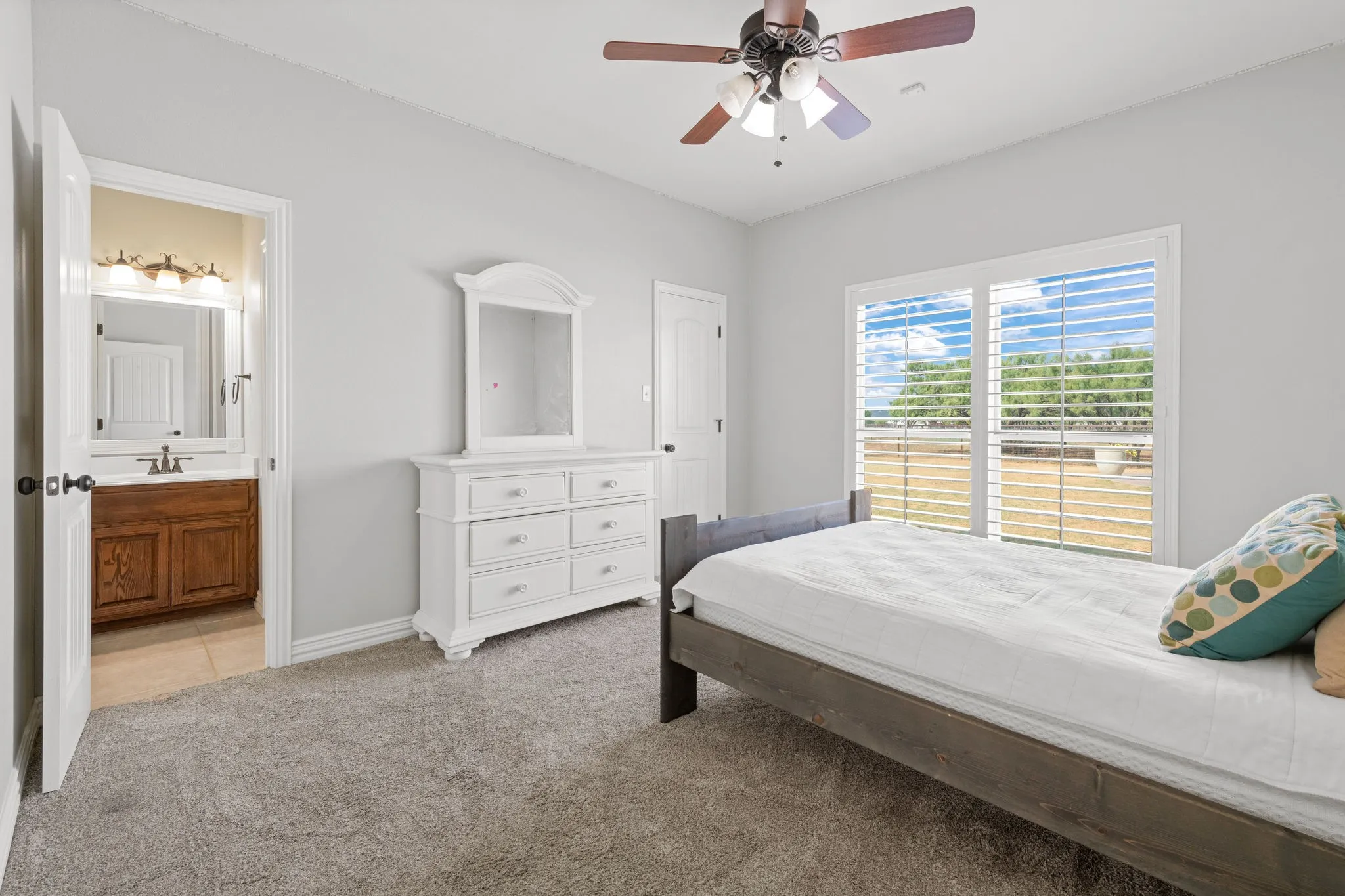 Carpeted room with a ceiling fan, a double window with wooden shutters, and an adjoining bathroom featuring a wood-finish vanity with a single sink.  Walk in closet.