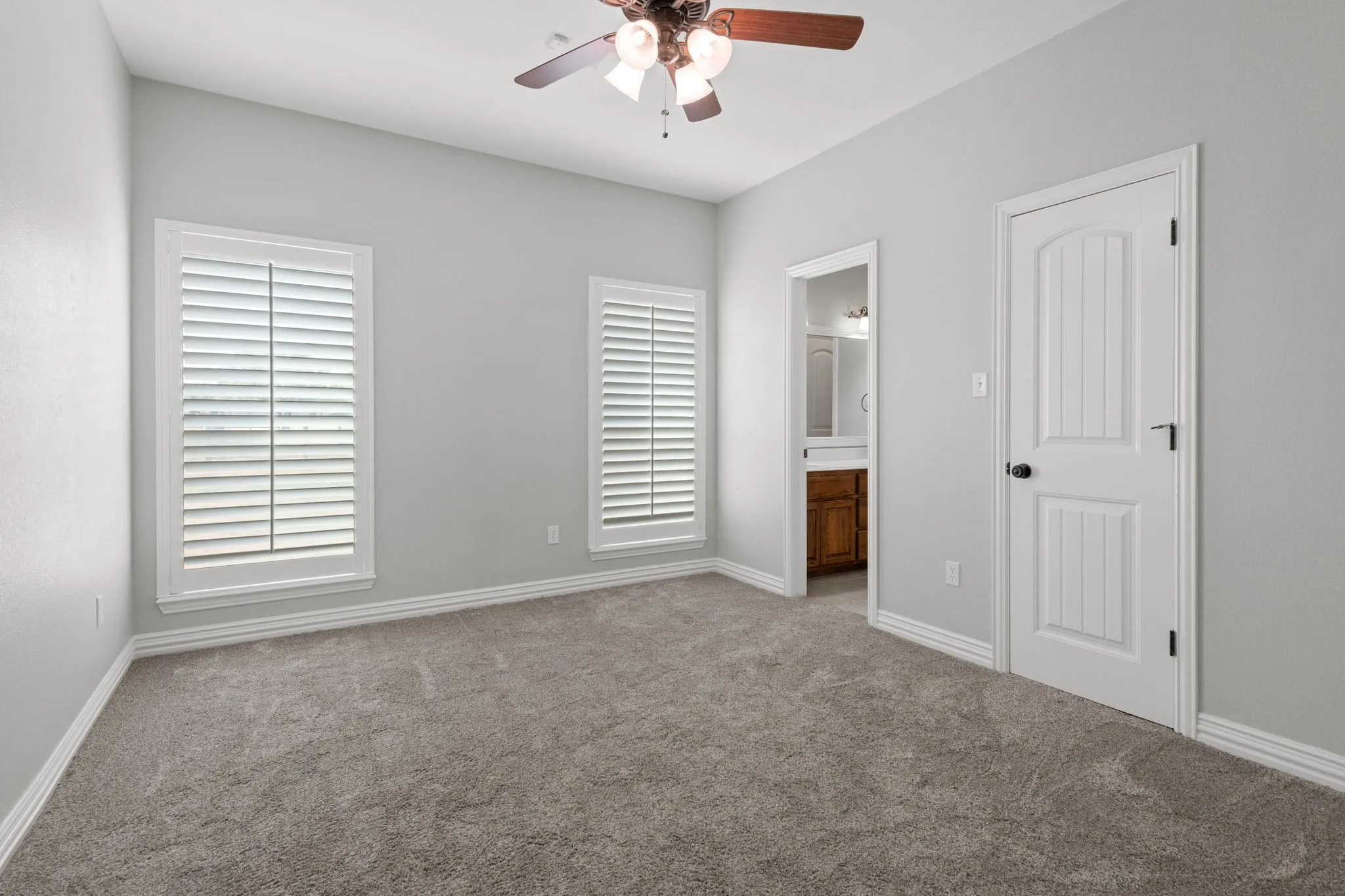 Carpeted room featuring light gray walls, two windows with white wood shutters, a wood-finish ceiling fan, and adjoining bathroom.
