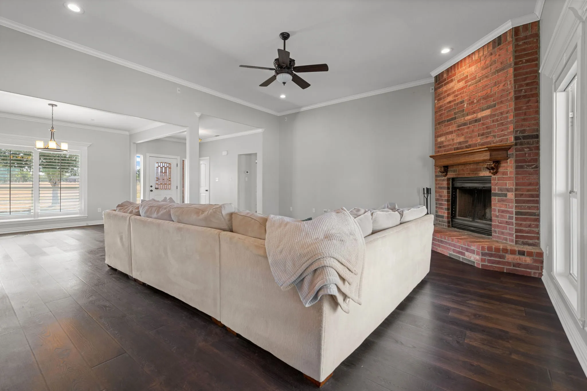 Spacious living area featuring dark wood-finish LVP flooring, a brick fireplace with a wood mantle, white crown molding, and recessed lighting.