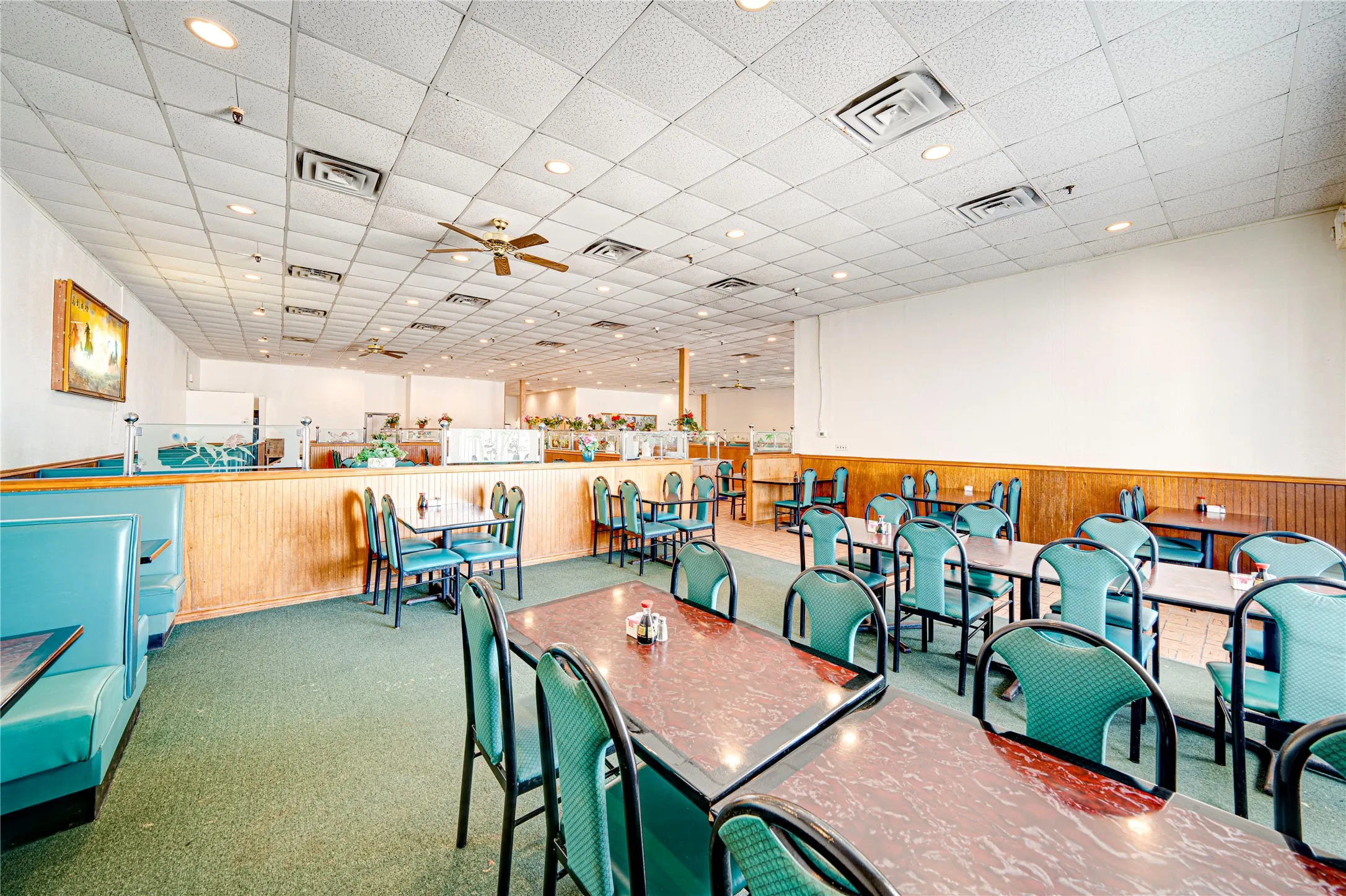 Spacious commercial dining area featuring a wood-finish service counter, green metal-frame seating throughout, wood-paneling wainscoting, drop ceiling with recessed and pendant lighting, and polished concrete flooring