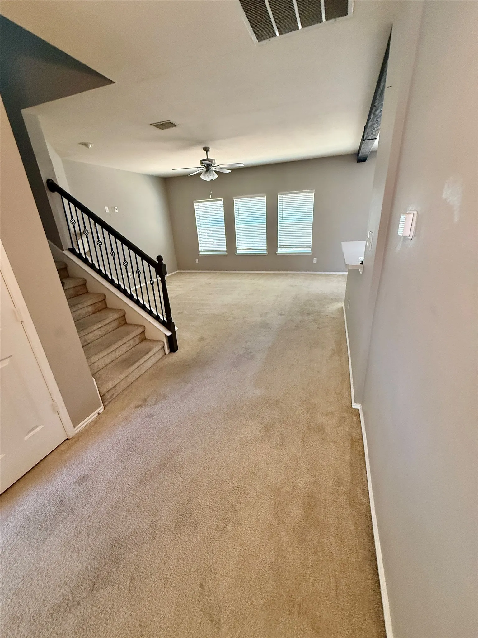 Two-story interior with beige carpeting, a staircase with black wrought iron balusters, a ceiling fan, and multiple windows with blinds