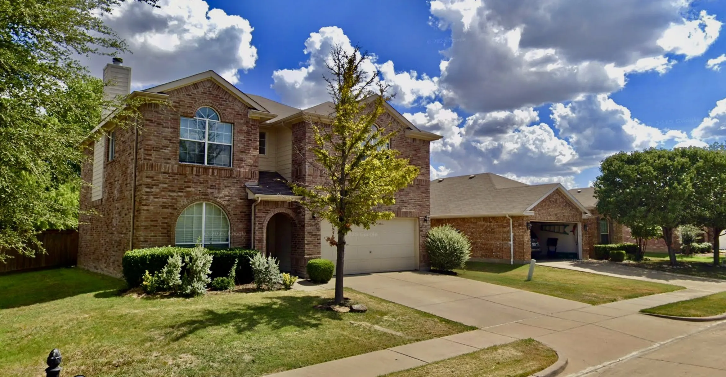 Brick facade with arched entryway, curved windows, and a two-car garage and mature landscaping