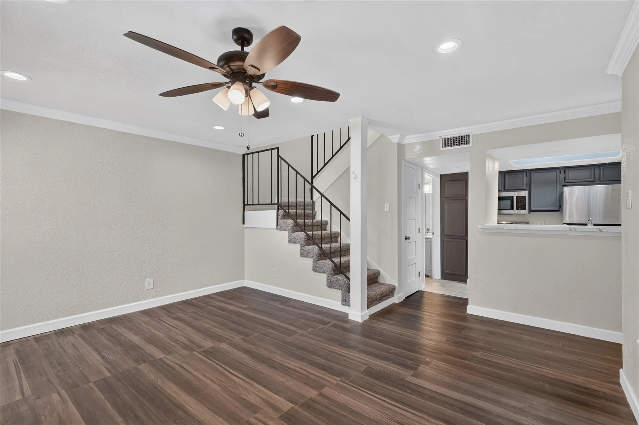 Wood-finish flooring, staircase with wrought iron railing, contemporary ceiling fan, recessed lighting, and kitchen pass-through with dark cabinetry