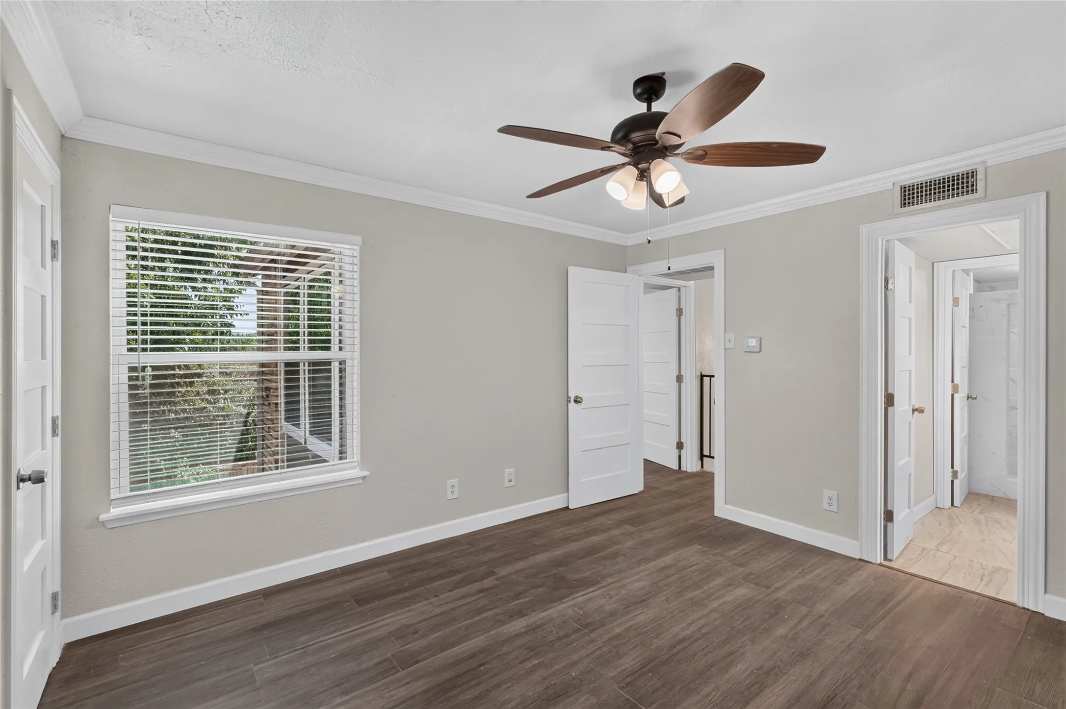 Spacious bedroom featuring dark wood-finish flooring, ceiling fan with integrated lighting, large double-hung window with plantation shutters, and multiple closet entries
