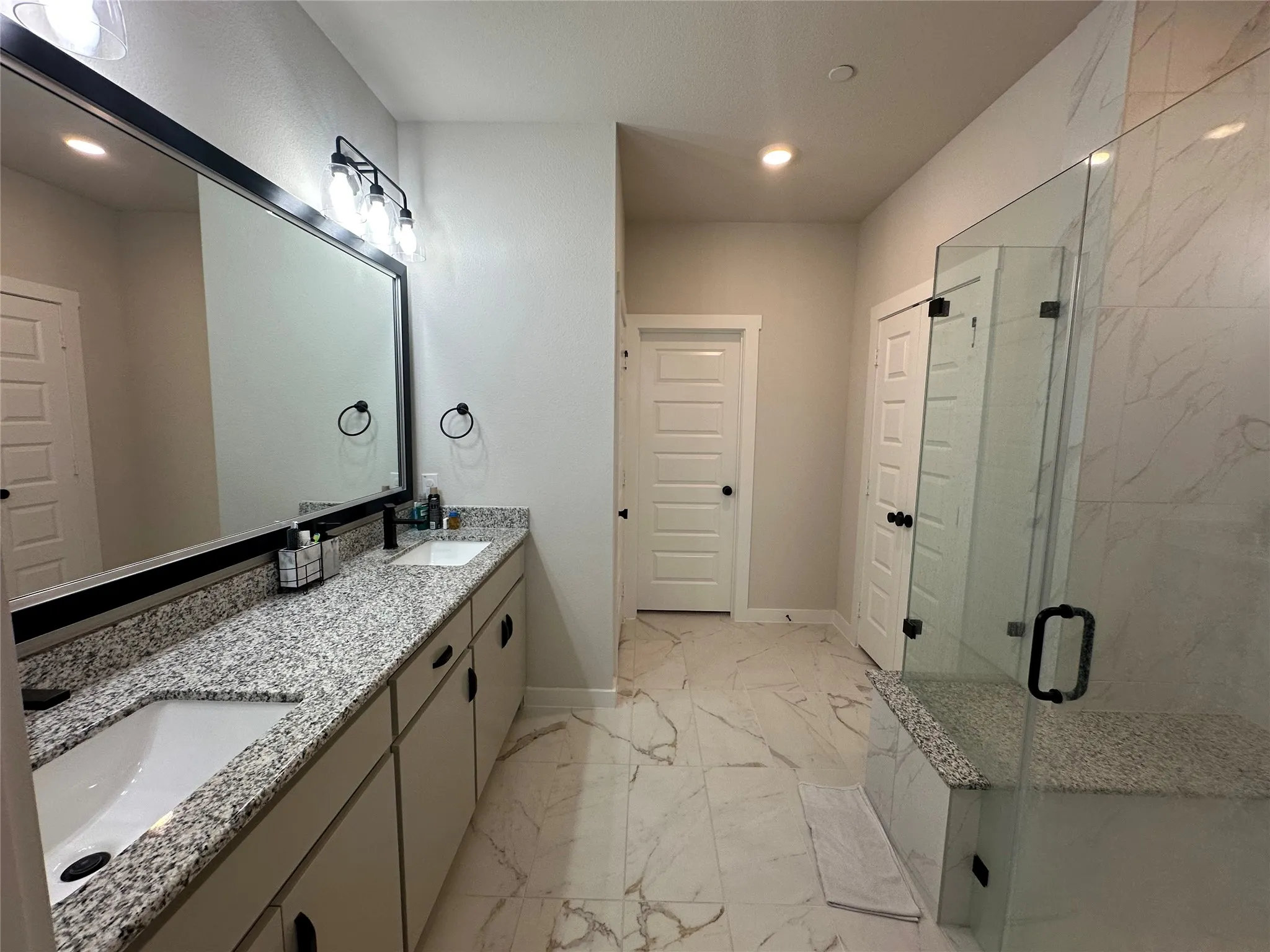 Spa-inspired primary bathroom featuring dual vanities with speckled quartz countertops, a frameless glass-enclosed shower with marble-look tile surround, and recessed ceiling lighting
