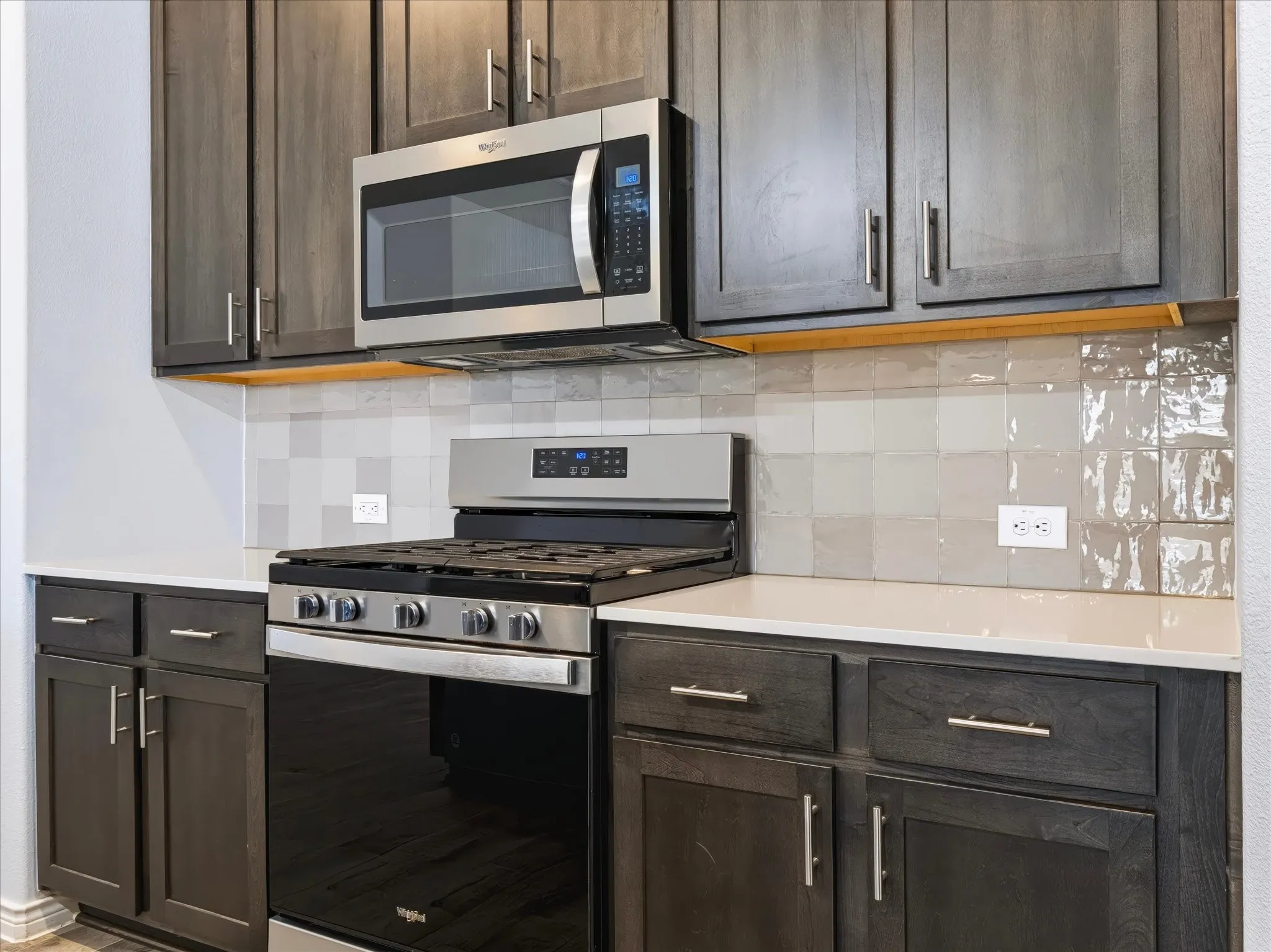 Gray wood-finish cabinetry, stainless steel microwave, stainless steel range, light-toned countertop, and a tiled backsplash