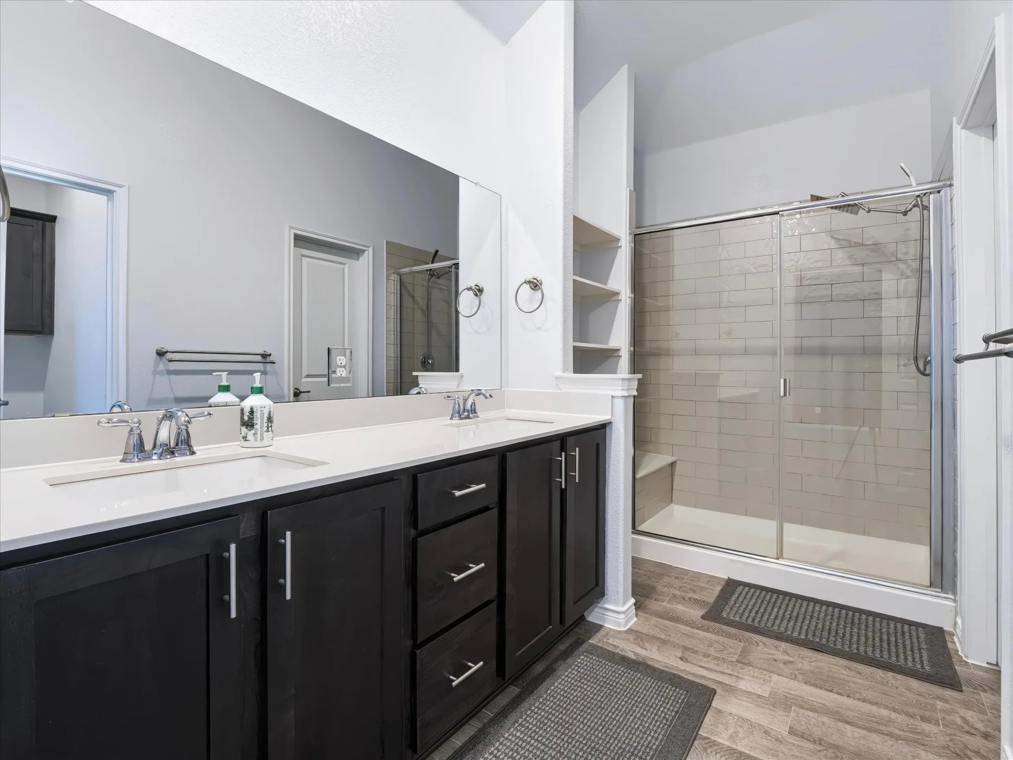 Dual vanity with integrated sinks and chrome fixtures, dark wood-finish cabinetry, built-in shelving, tiled shower with glass enclosure, and wood-finish flooring