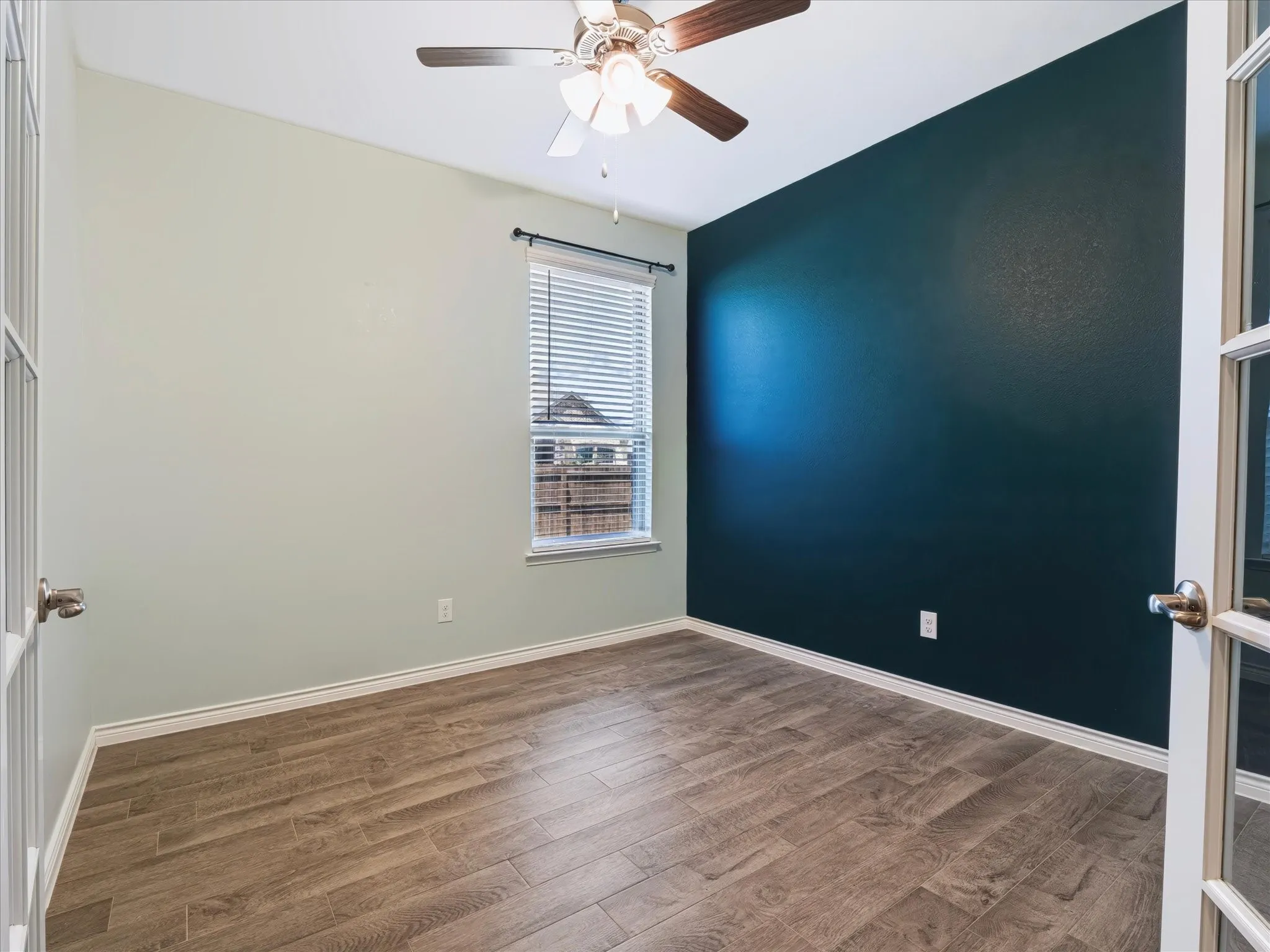 Wood-finish flooring with white baseboards, a window with horizontal blinds, a ceiling fan with integrated lighting, and French doors