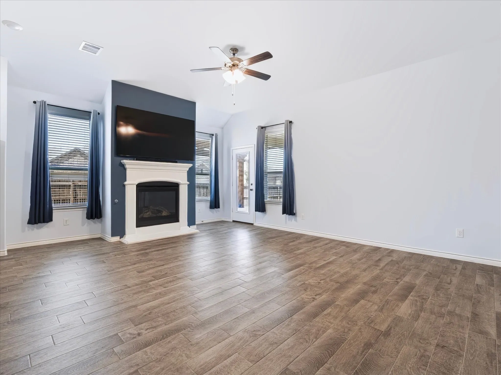 Spacious room featuring wood-finish flooring, a decorative fireplace with a mantel, a ceiling fan, and multiple windows with blinds