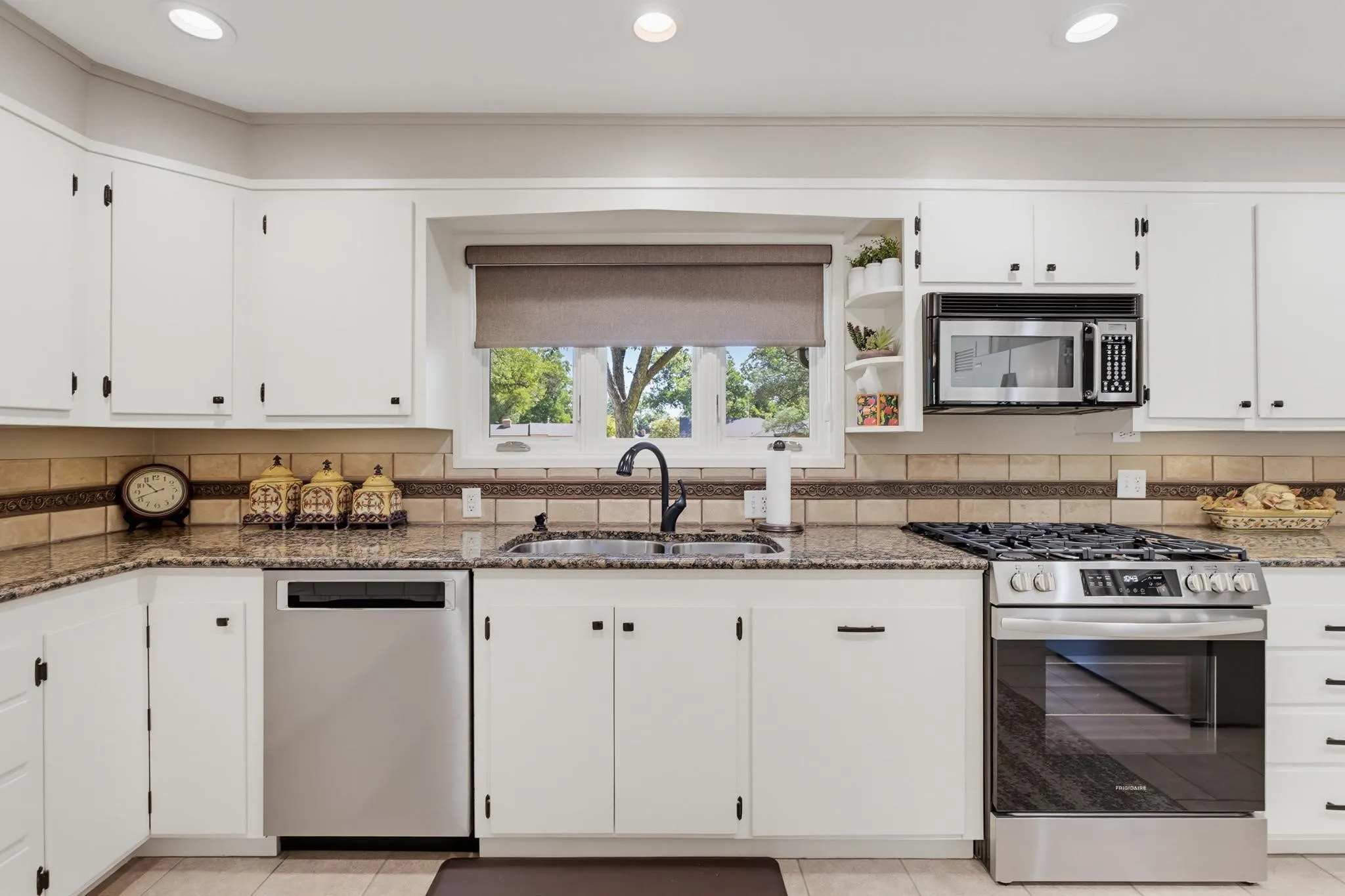 White cabinetry with black hardware, stone countertops, tiled backsplash, stainless steel appliances, and a window above the sink