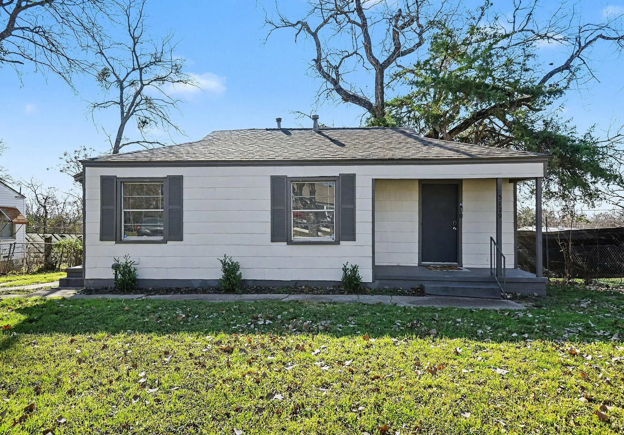 White exterior siding with dark shutters, covered entry porch, dark entry door, and a front yard with mature trees