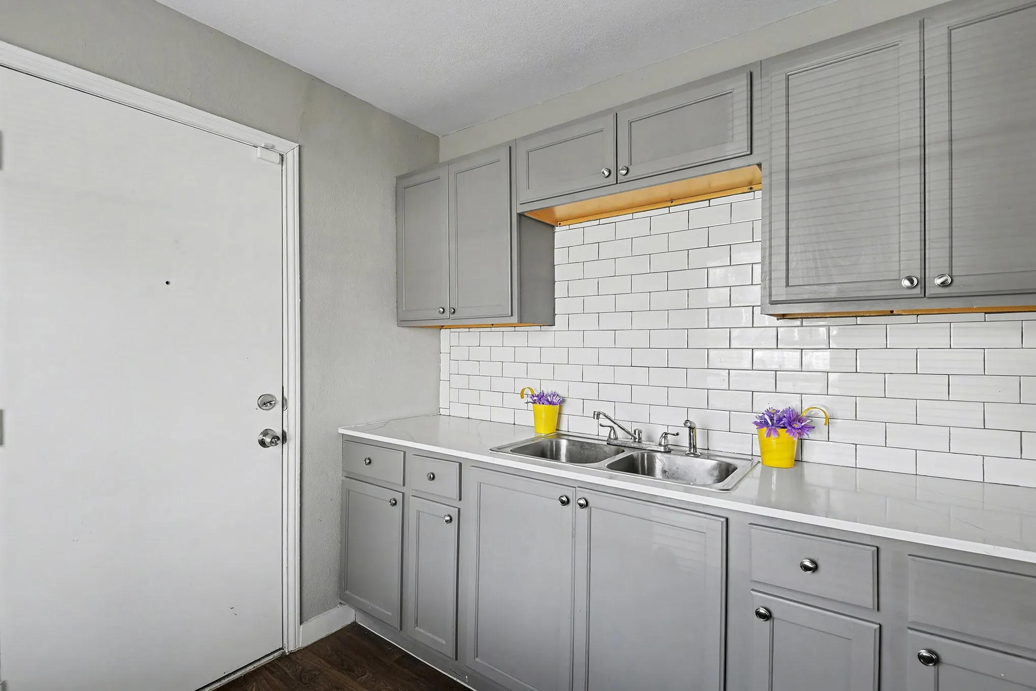 Gray cabinetry with silver hardware, white subway tile backsplash, light-colored countertops, and a double basin sink
