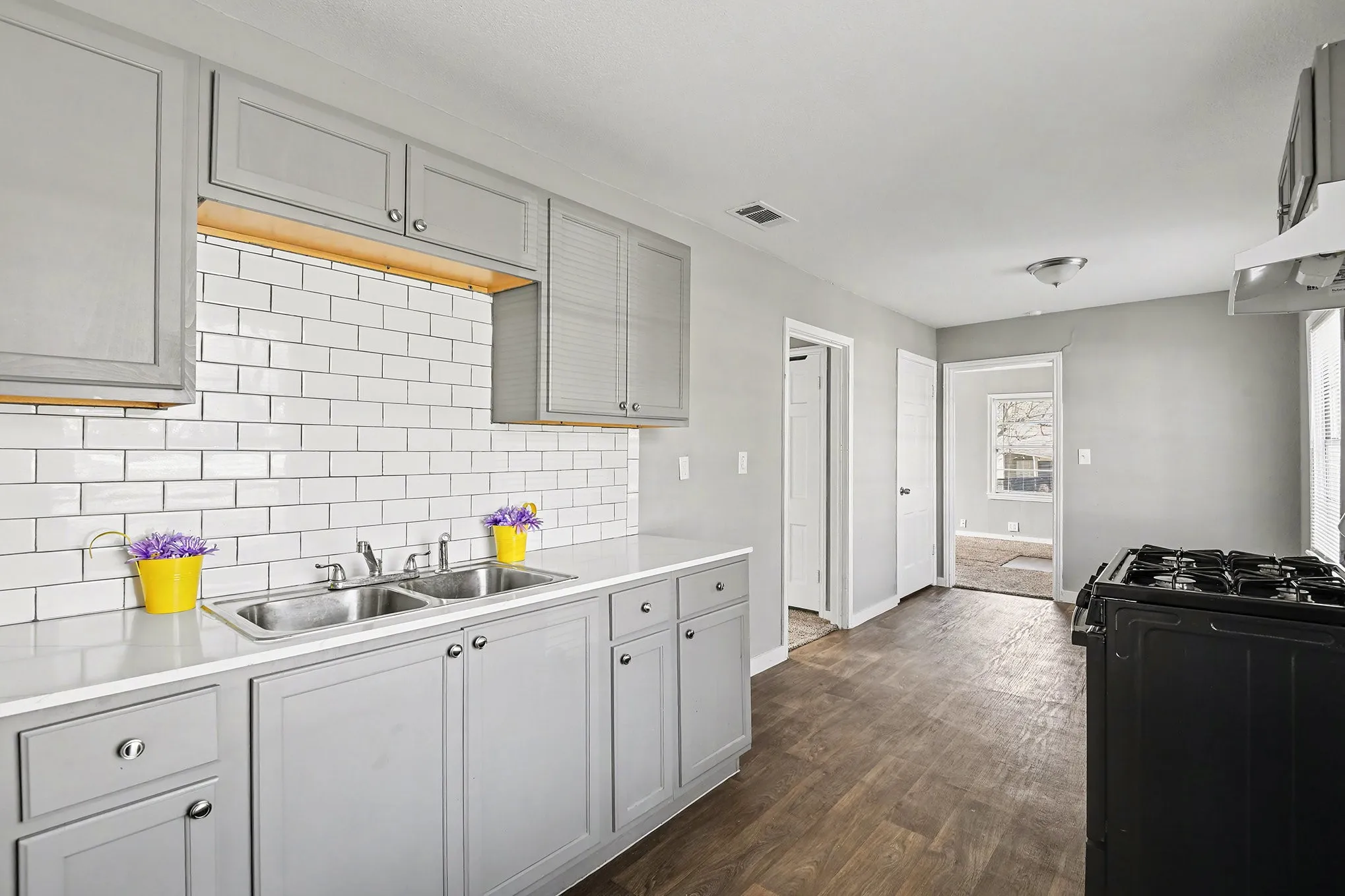 Kitchen featuring gray shaker-style cabinetry, white subway tile backsplash, a double basin stainless steel sink, light-toned countertops, and wood-finish flooring