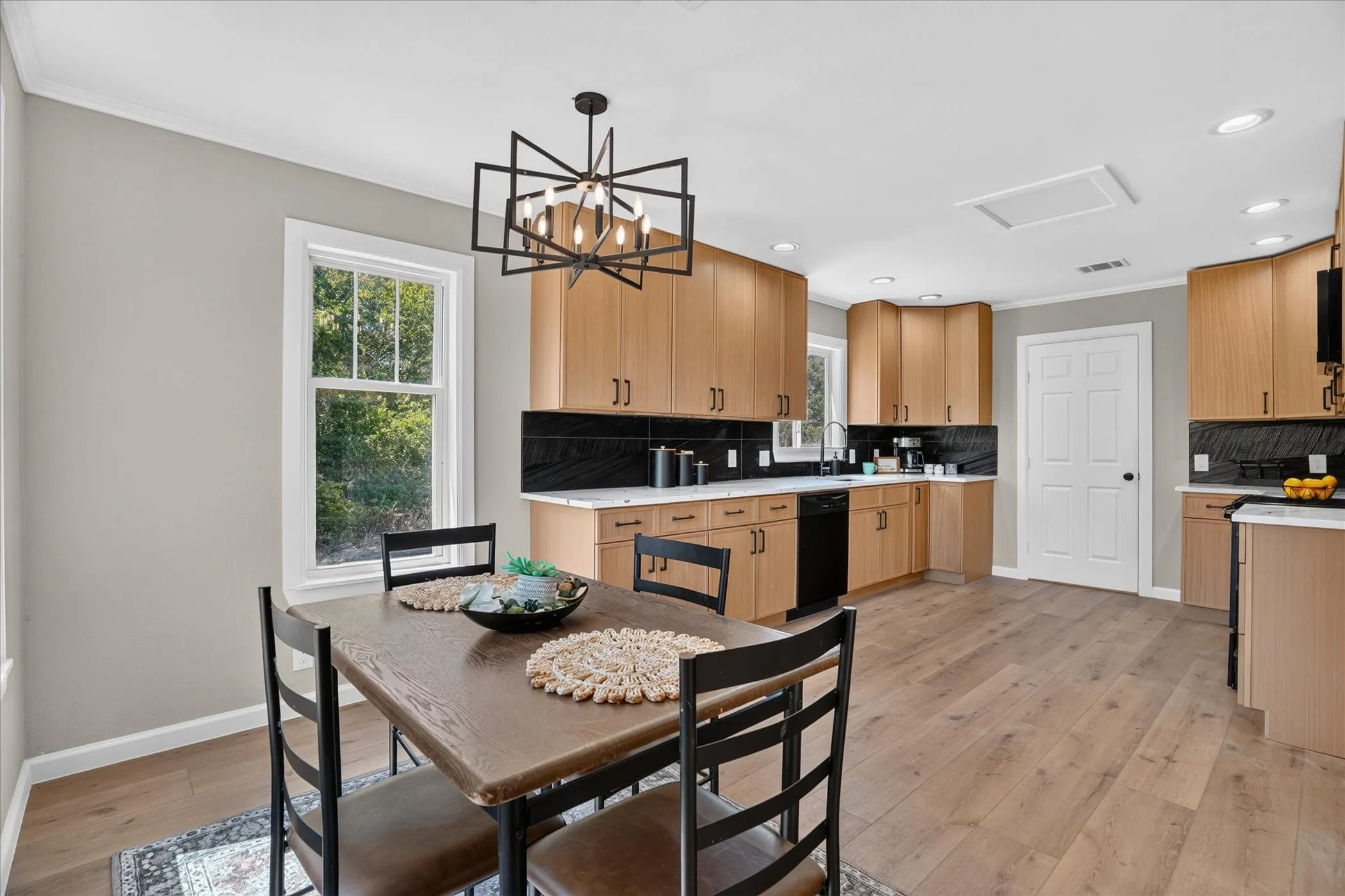 Open-concept kitchen with wood-finish flooring, light wood cabinetry, black subway tile backsplash, and white countertops