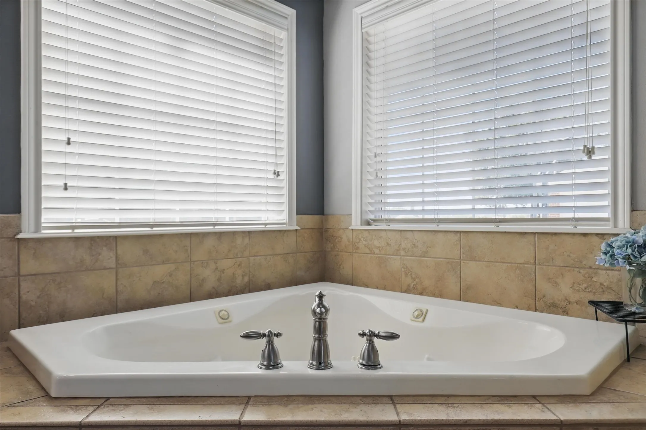 Corner soaking tub with polished chrome fixtures, surrounded by neutral-toned tile and two windows featuring white horizontal blinds