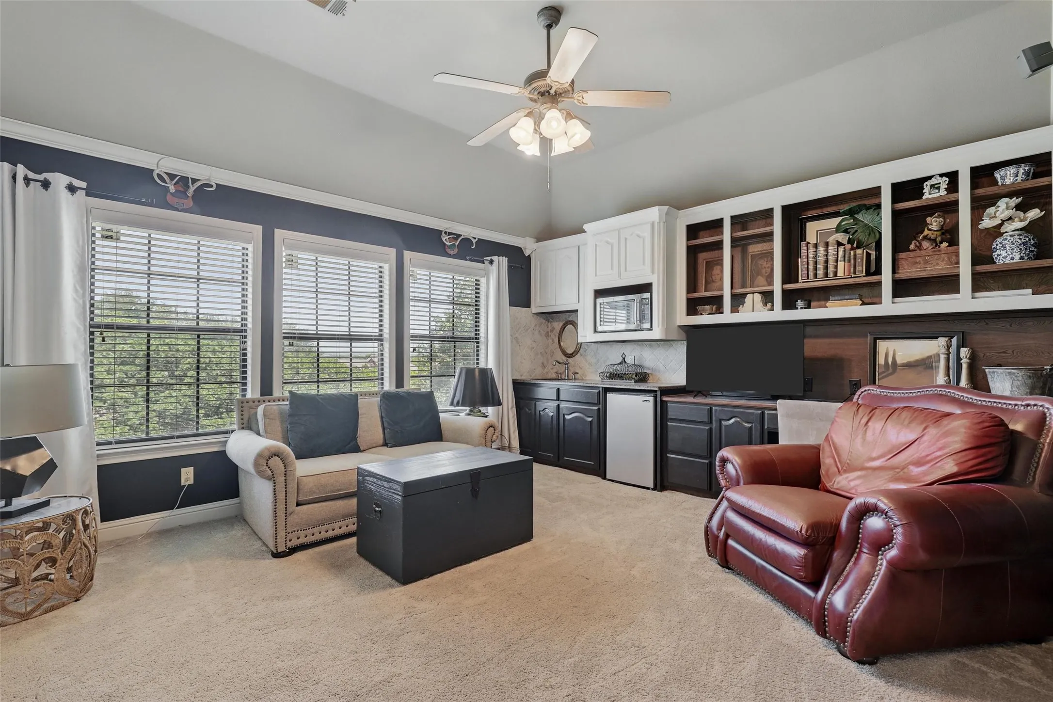 Carpeted room featuring multiple windows with blinds, a built-in wet bar with white upper cabinetry and dark lower cabinetry, a built-in microwave, and open shelving