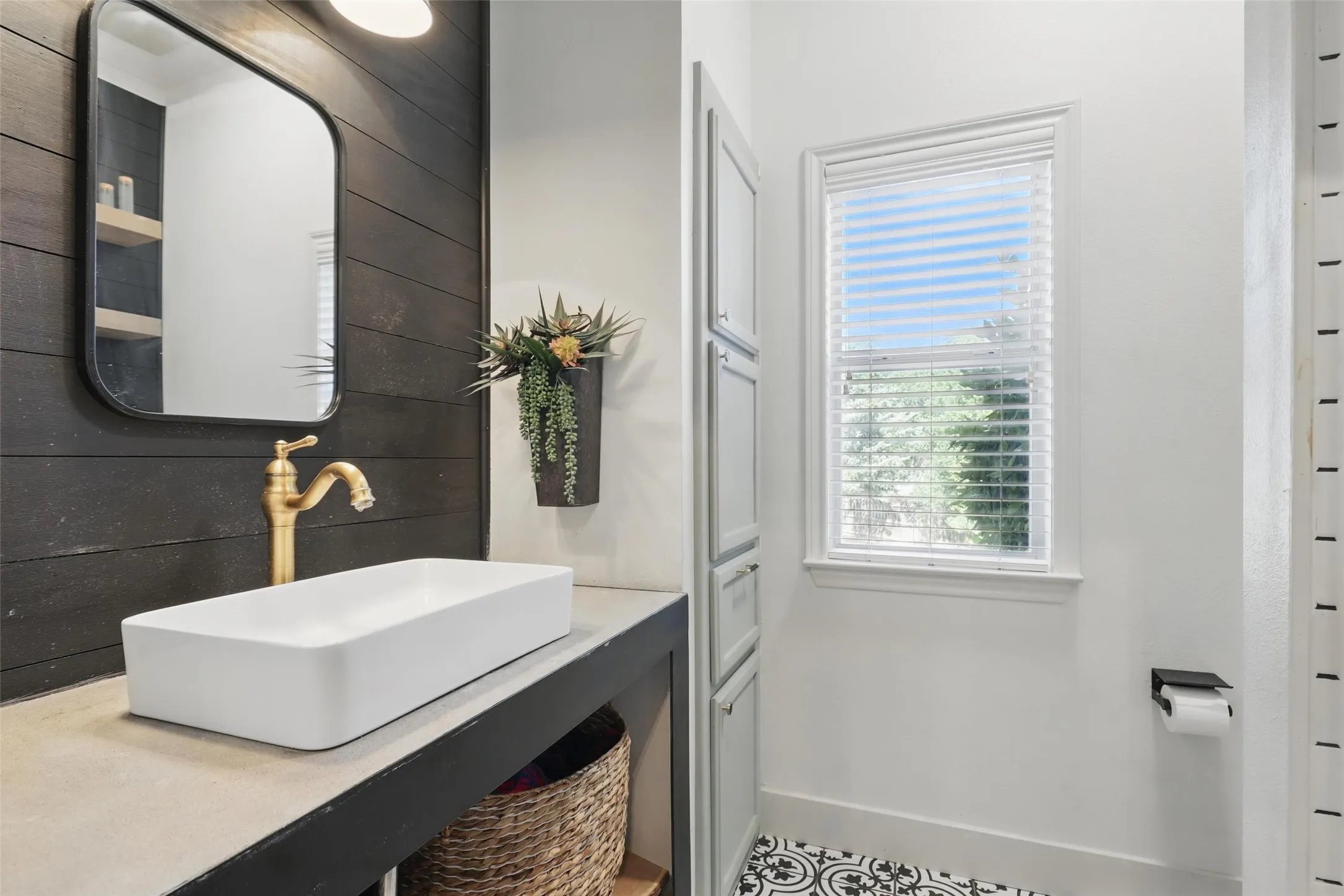 Bathroom featuring a vessel sink with a brass-finish faucet, dark wood paneling, built-in storage cabinetry, patterned floor tiling, and window with blinds
