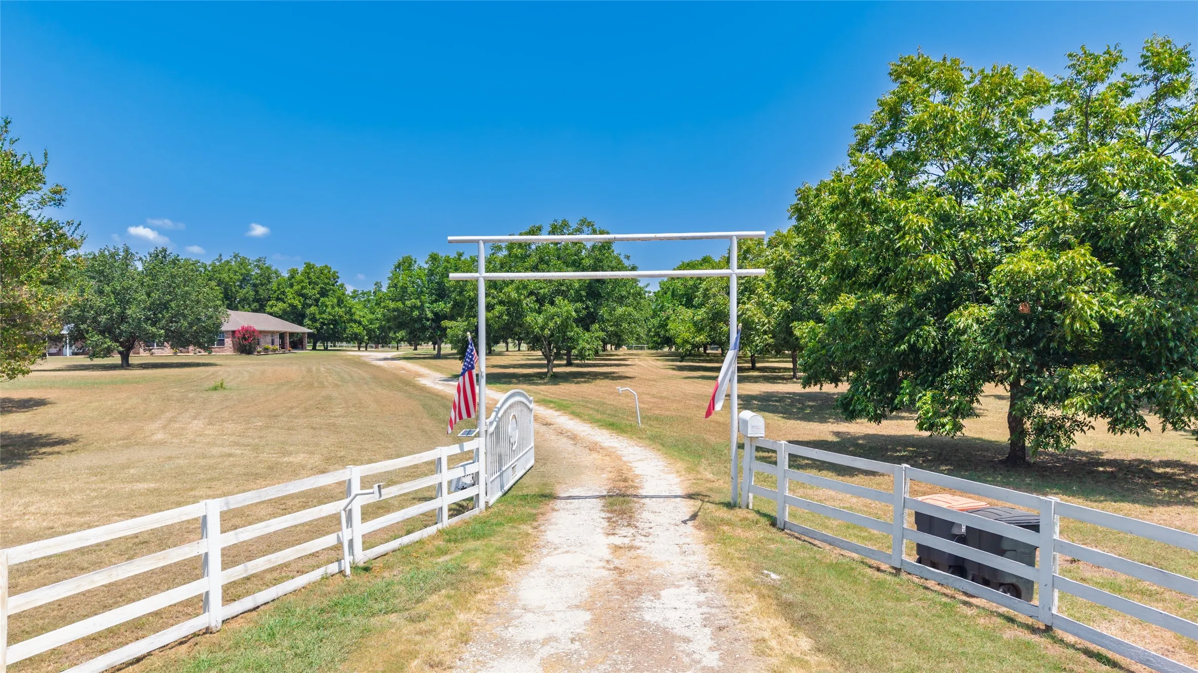 Gated entry with a white rail fence, a long gravel driveway, and mature trees