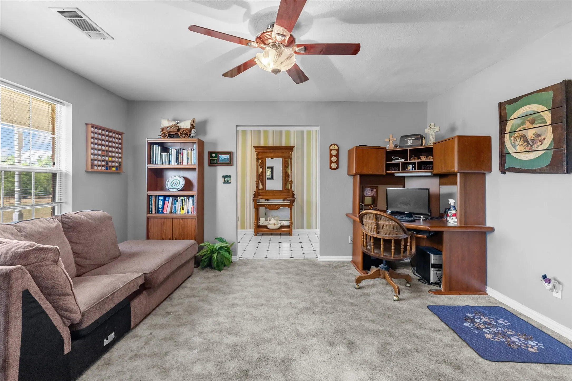 Spacious recreation room featuring neutral carpeting, built-in wood cabinetry with shelving and desk workspace, ceiling fan with light fixture, large windows with natural light, and hardwood accent furniture pieces throughout the open layout