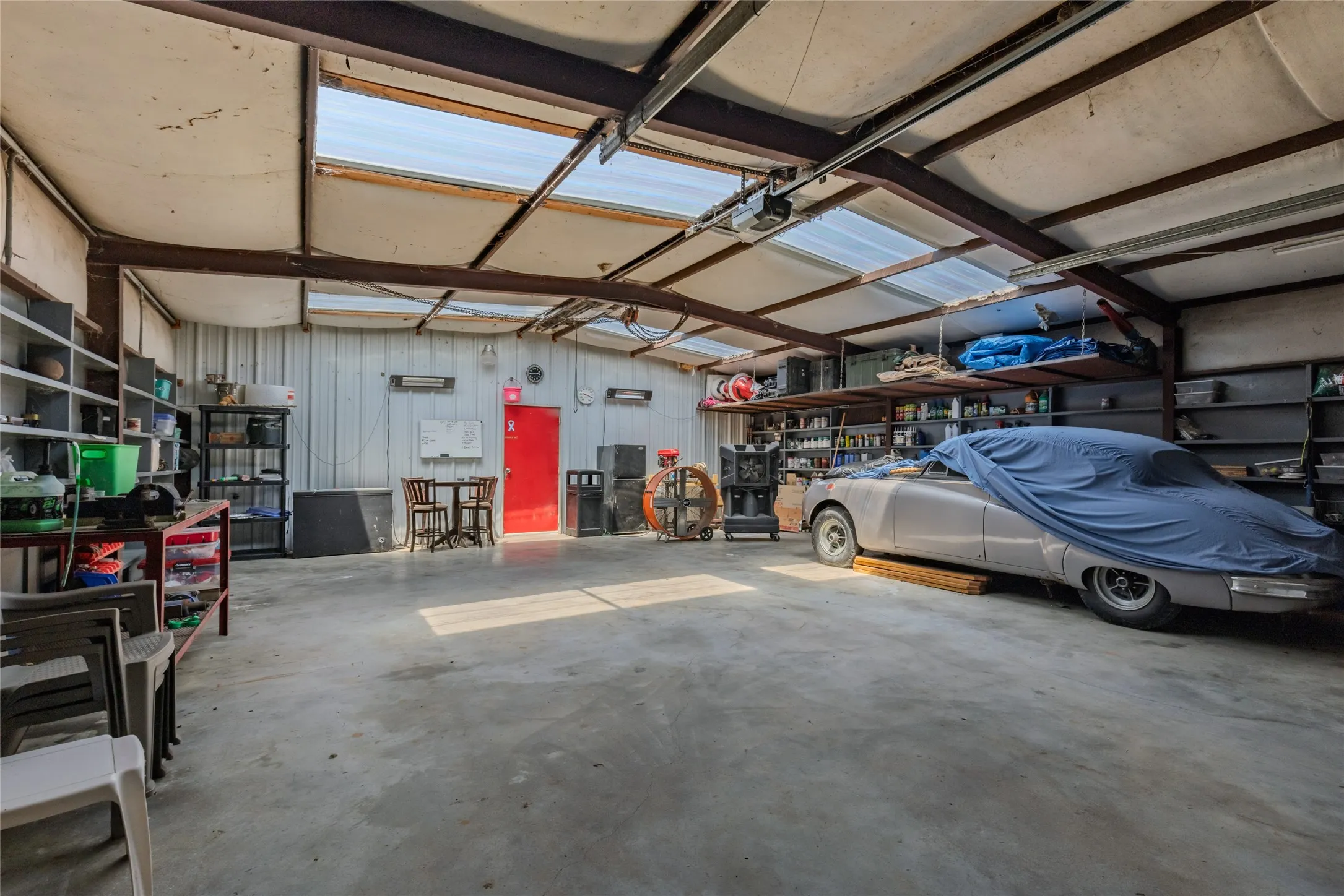 Metal-framed structure with a concrete floor, equipped with overhead skylights, built-in shelving, and a red entry door