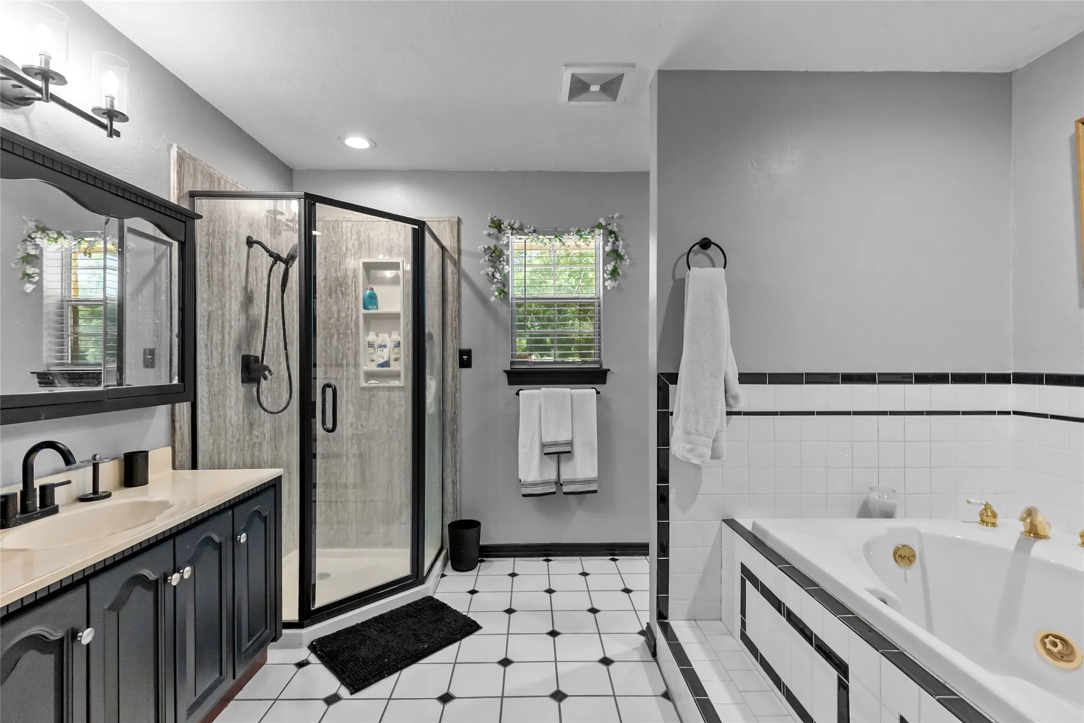 Built-in soaking tub, separate shower enclosure with glass surround, black and white tiled flooring, single vanity with integrated sink, and wall-mounted light fixture