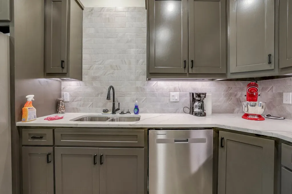 Dark gray cabinetry with black hardware, undermount double-basin stainless steel sink, black gooseneck faucet, light-toned stone countertop, and subway tile backsplash