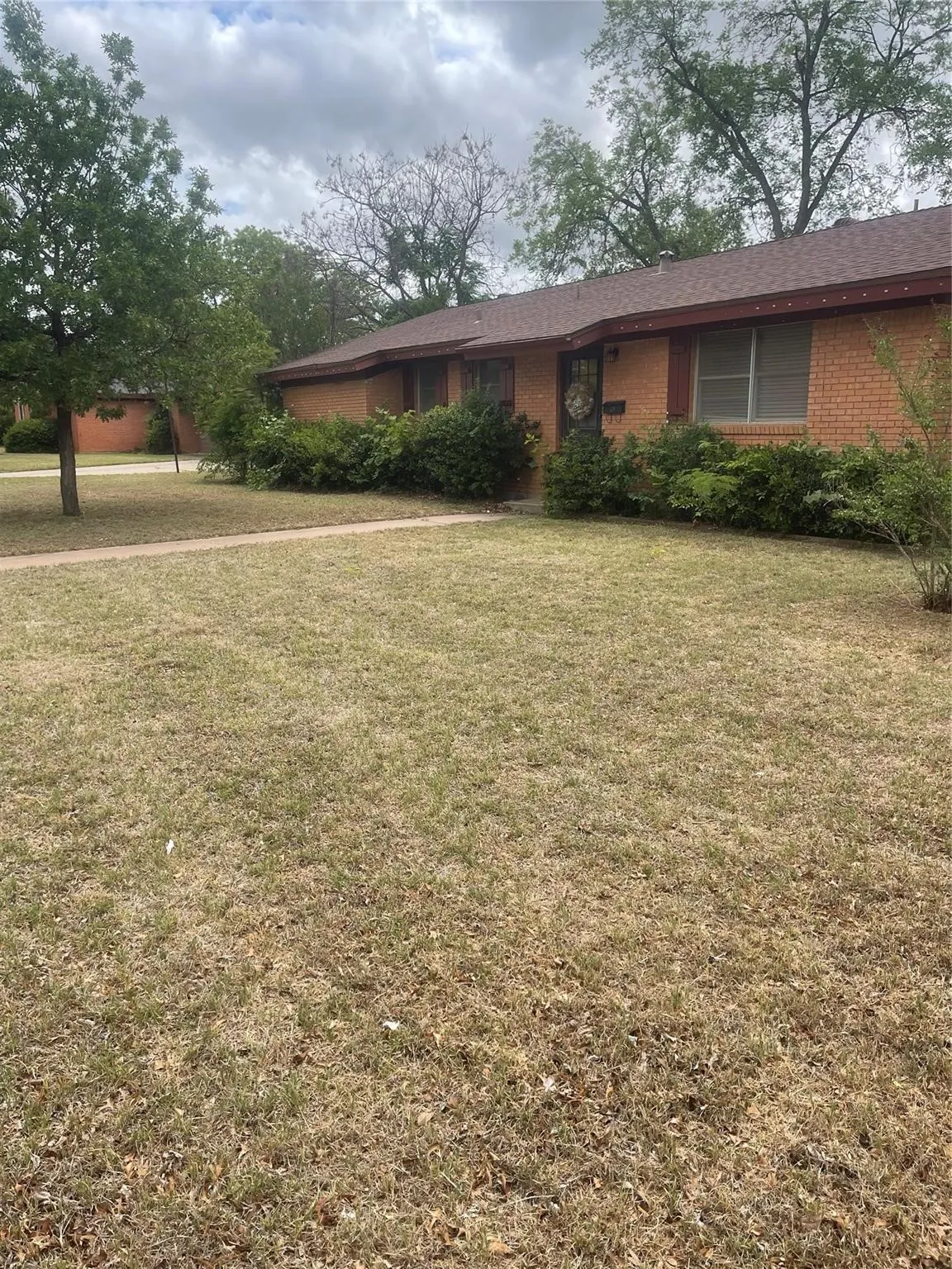 Brick exterior with a brown shingle roof, established landscaping, and a concrete walkway