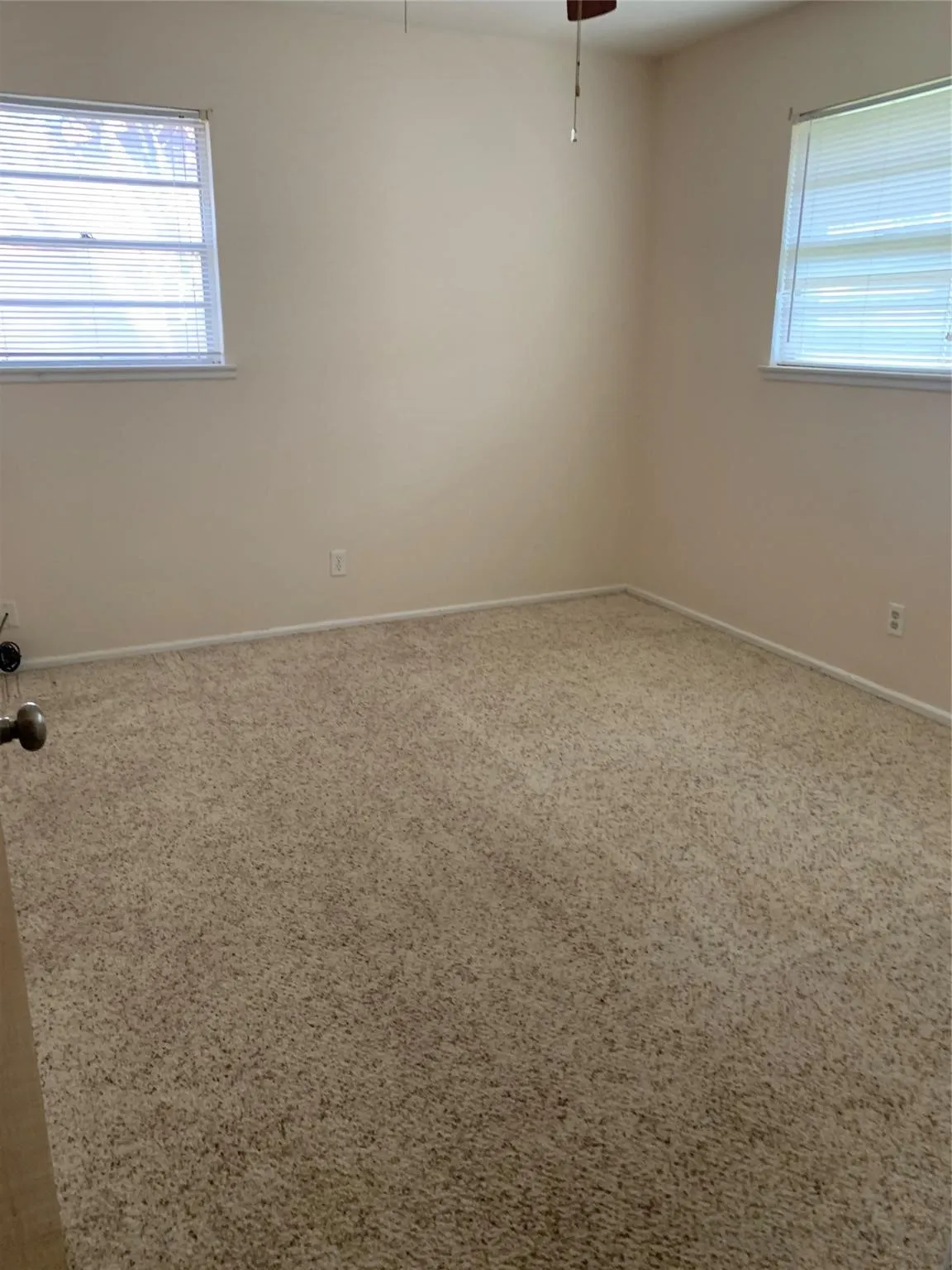 Plush carpet flooring with neutral wall tones, two windows featuring horizontal blinds, and a ceiling-mounted fan
