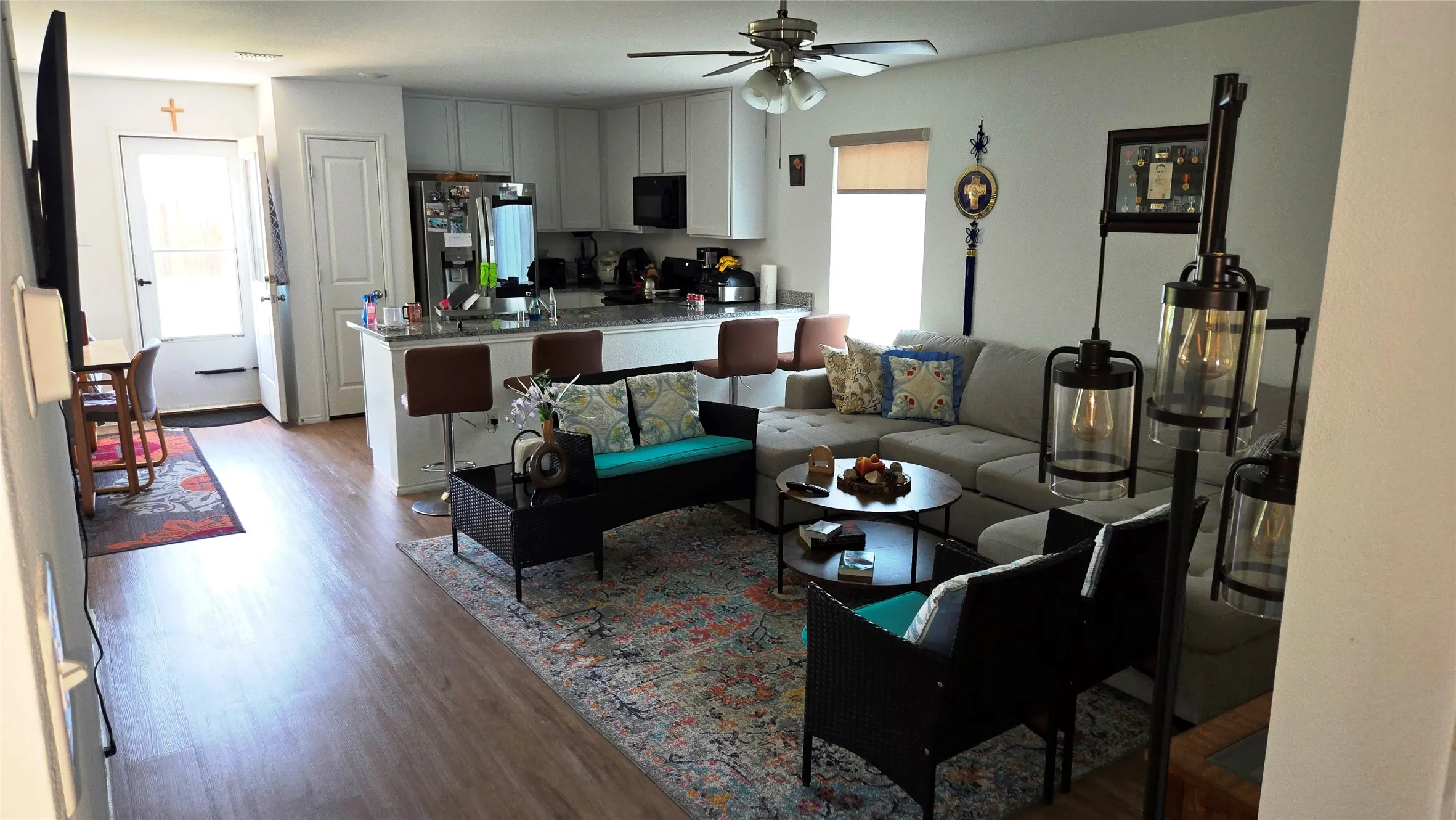Open-concept living area featuring wood-finish flooring, a kitchen island with seating, upper and lower white cabinetry, a built-in microwave, and a ceiling fan