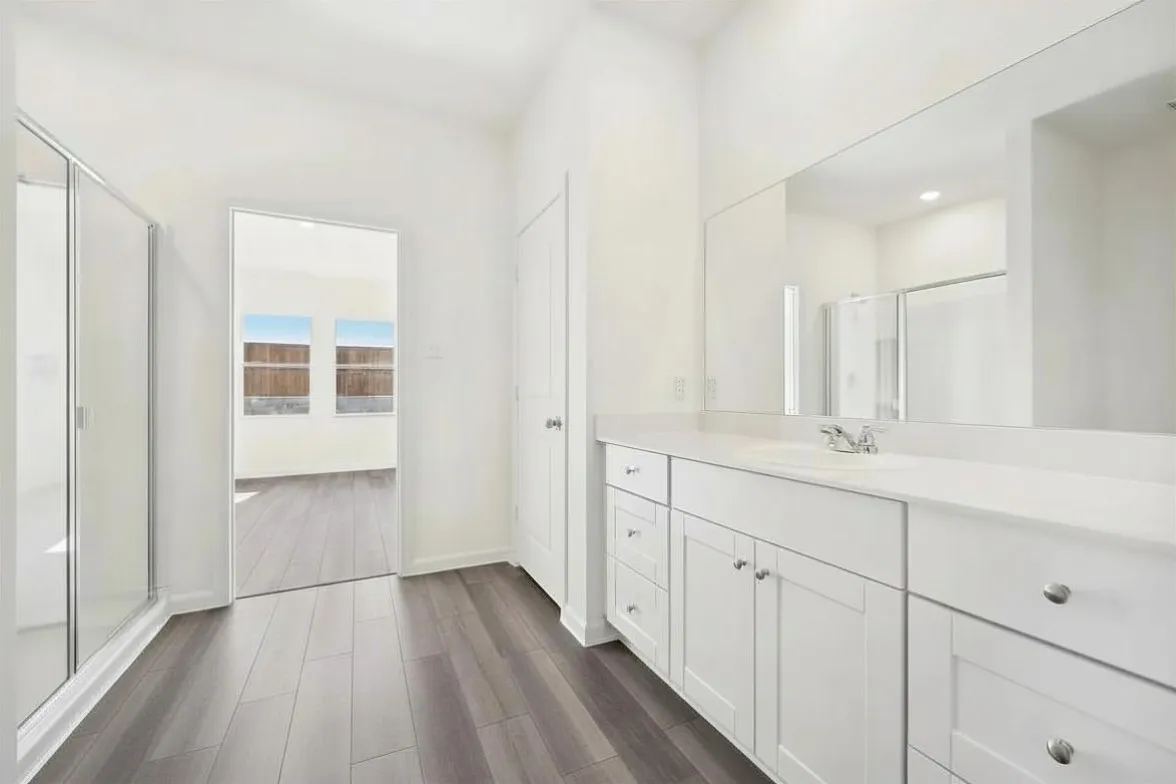 Spacious bathroom featuring white cabinetry, a light-colored countertop, dual sinks, a large wall-mounted mirror, a glass-enclosed shower, and wood-finish flooring