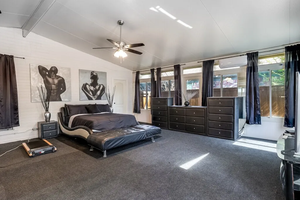 Vaulted ceiling with exposed beams, ceiling fan, white painted brick wall, multiple transom windows, and wall-mounted air conditioning unit