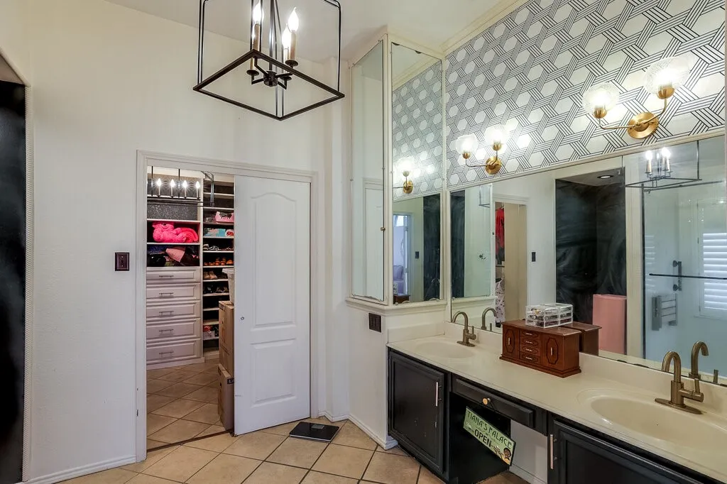 Dual vanity with ample counter space, custom patterned wall covering, decorative wall sconces, large framed mirror, and a walk-in closet with built-in storage