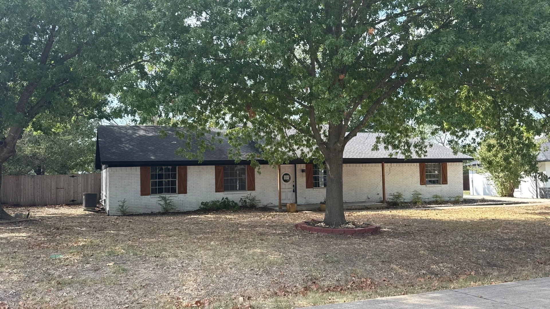 White brick exterior with a dark shingled roof, featuring wood-finish shutters and a partially fenced yard