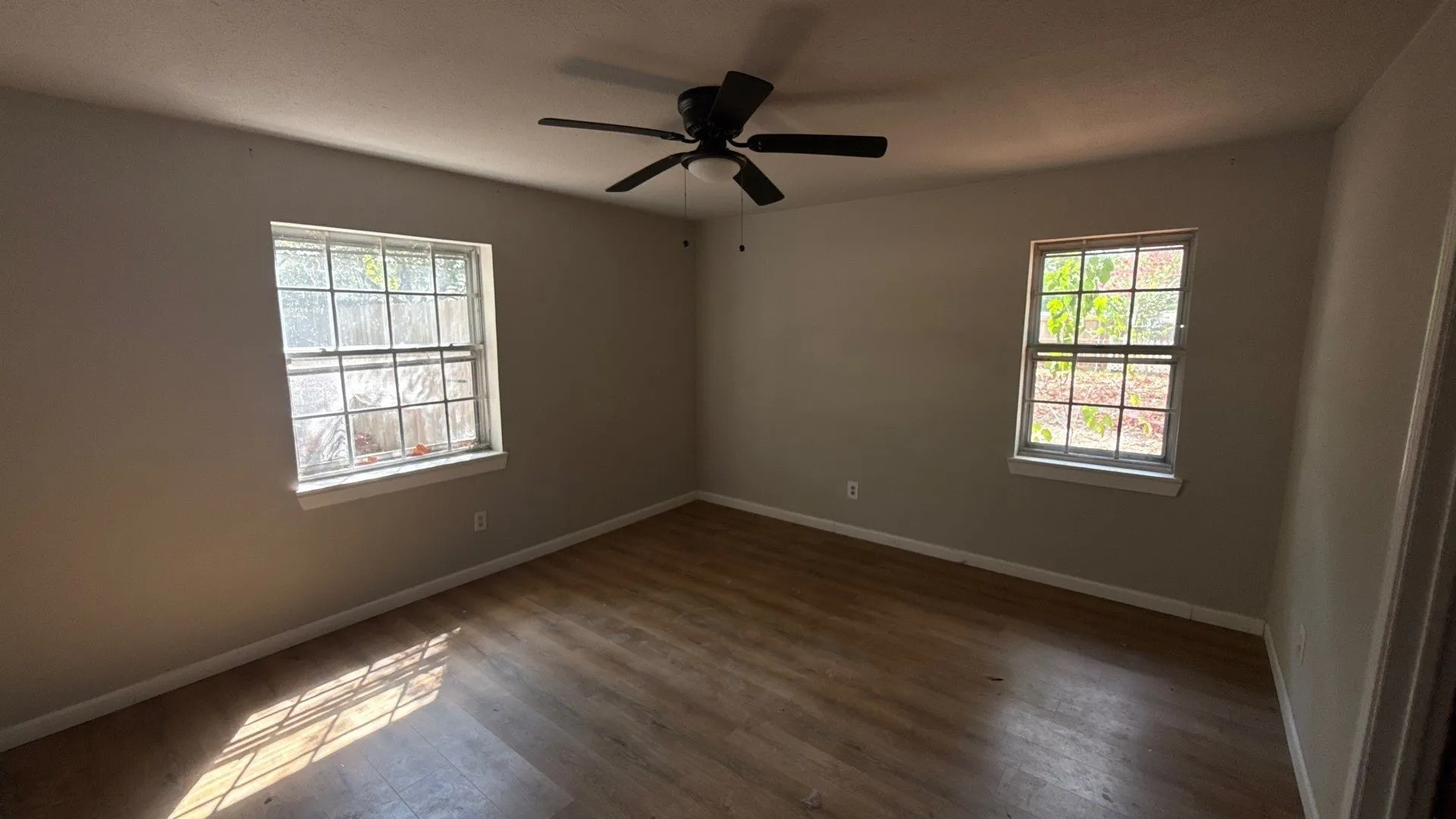 Wood-finish flooring, two large windows, neutral wall tones, and a ceiling fan with integrated lighting