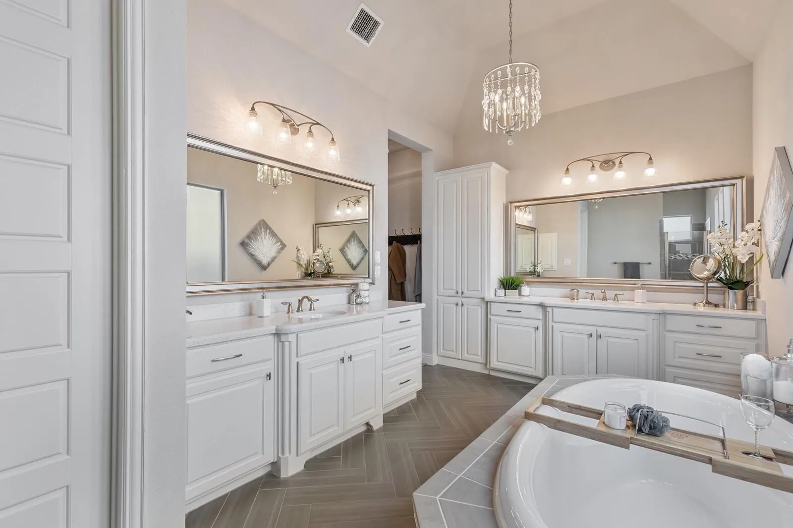 Vaulted ceiling with an elegant chandelier, dual vanity with undermount sinks, built-in cabinetry, large soaking tub, and herringbone-patterned floor tile