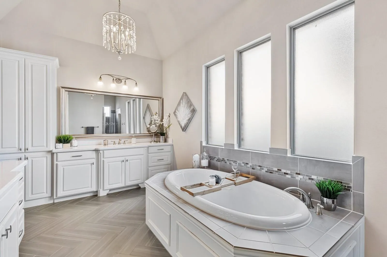 Oval soaking tub, white shaker cabinetry, herringbone-patterned wood-finish flooring, crystal chandelier, and frosted rectangular windows