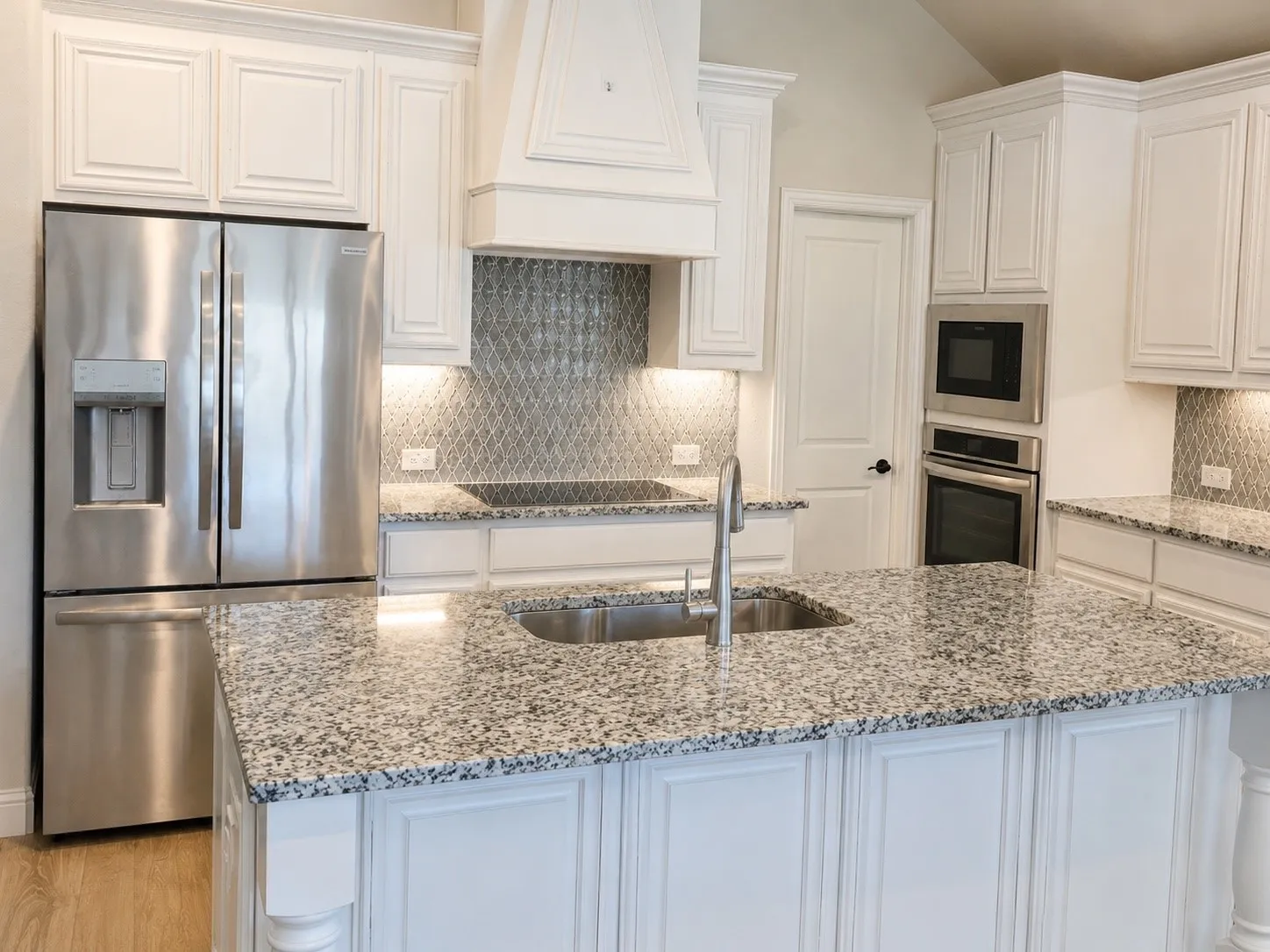 White cabinetry, stainless steel appliances, speckled countertops, kitchen island with sink, and a decorative tile backsplash