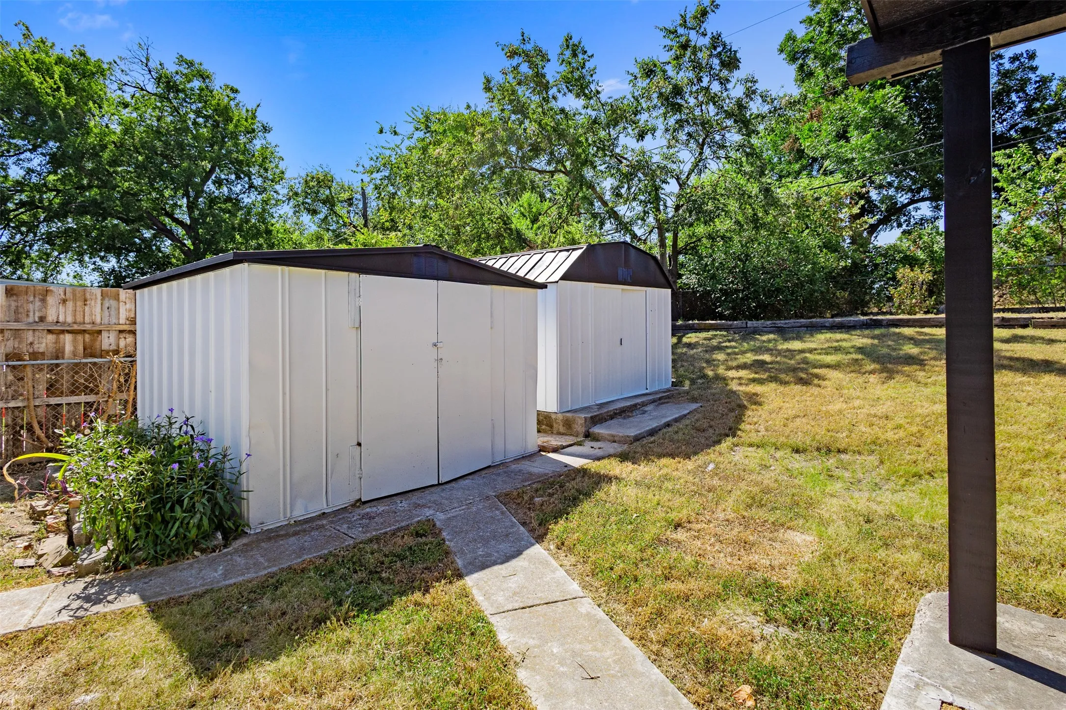 Two white storage sheds with dark roofs, concrete pads, and a concrete pathway