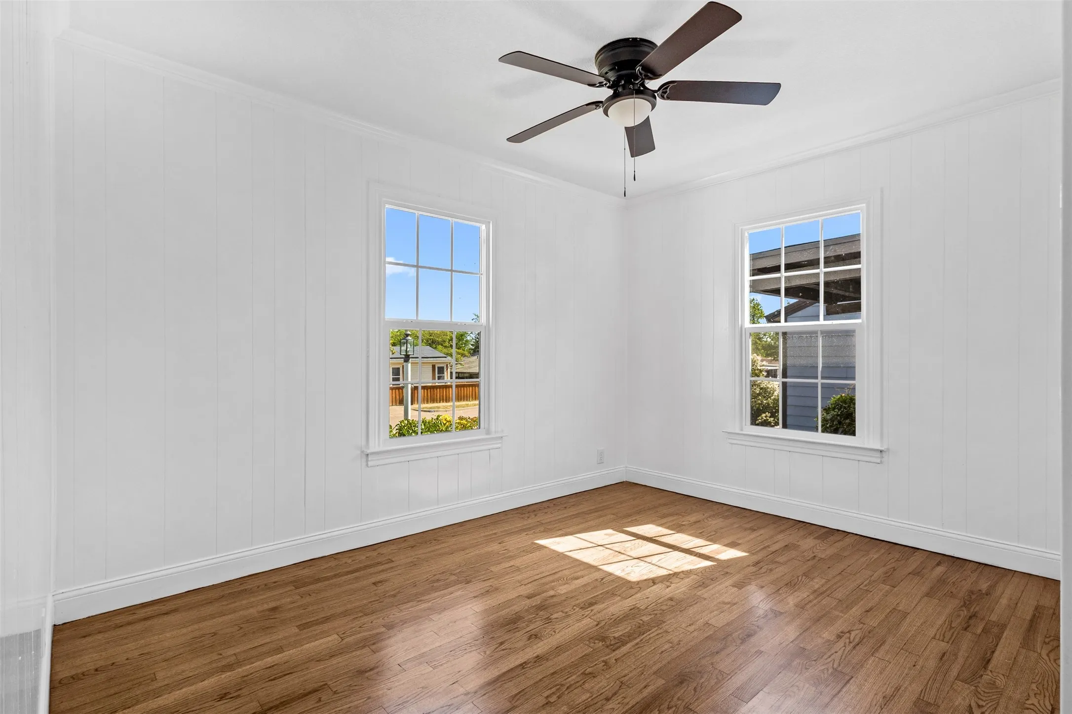 Hardwood flooring, two large windows, white paneling, ceiling fan with integrated lighting, and crown molding