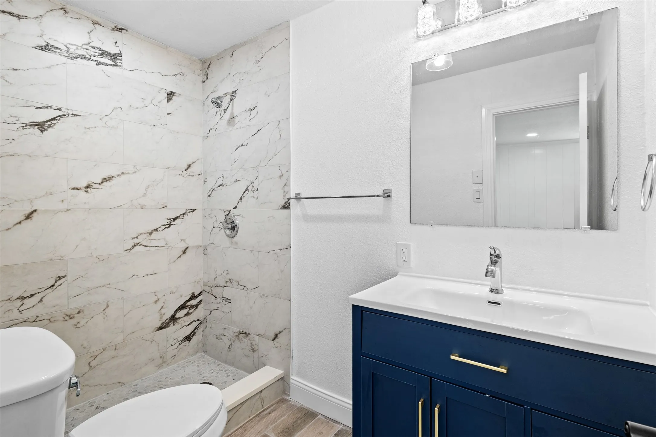 Walk-in shower with large format tile, a vanity with a deep blue finish, sleek gold-tone hardware, a white integrated sink, and an overhead vanity light fixture