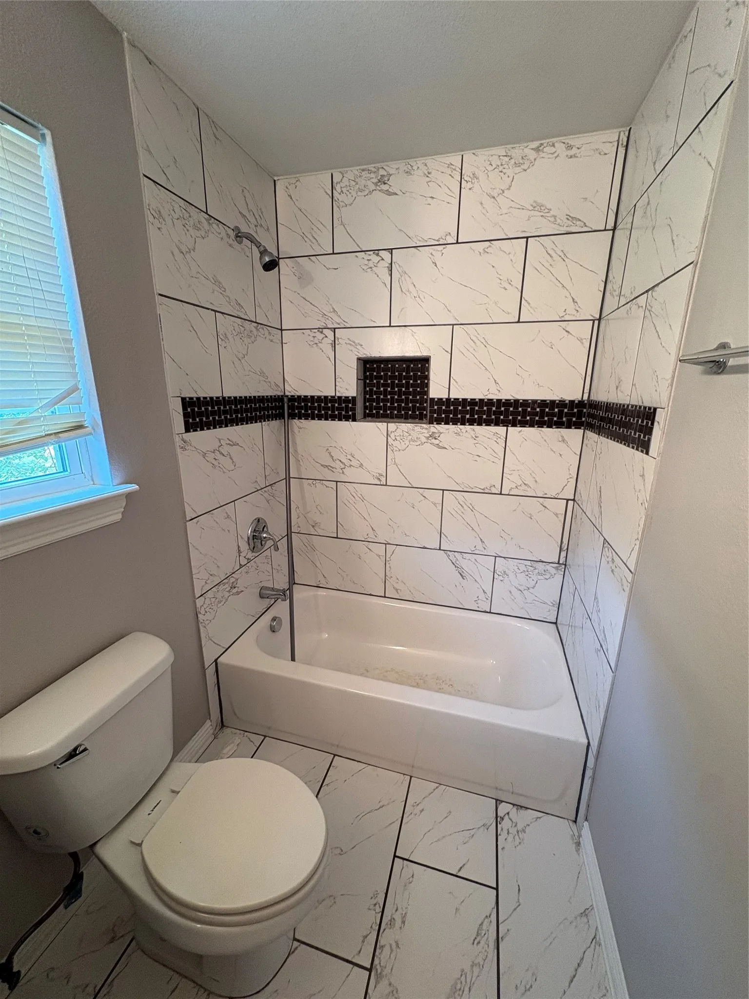Bathtub with a shower fixture, white and grey tiled surround, recessed shower niche, and a contrasting black accent tile band